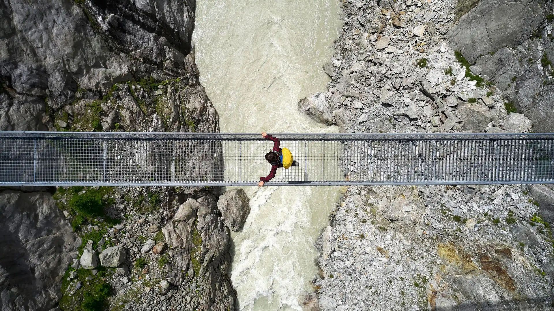 Persona con camicia rossa e zaino giallo attraversa ponte sospeso sopra fiume impetuoso in gola rocciosa