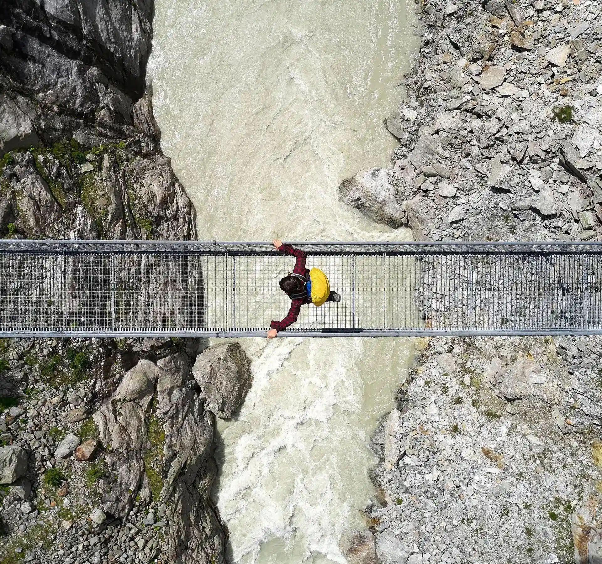 Person mit rotem Hemd und gelbem Rucksack überquert Hängebrücke über reissenden Fluss in felsiger Schlucht