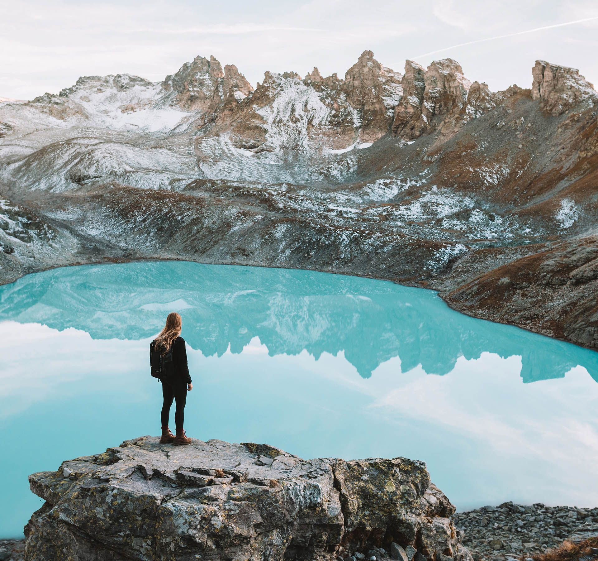 A woman stands on a rock in front of a turquoise blue lake of glacial water. Edged rock peaks can be seen in the background.