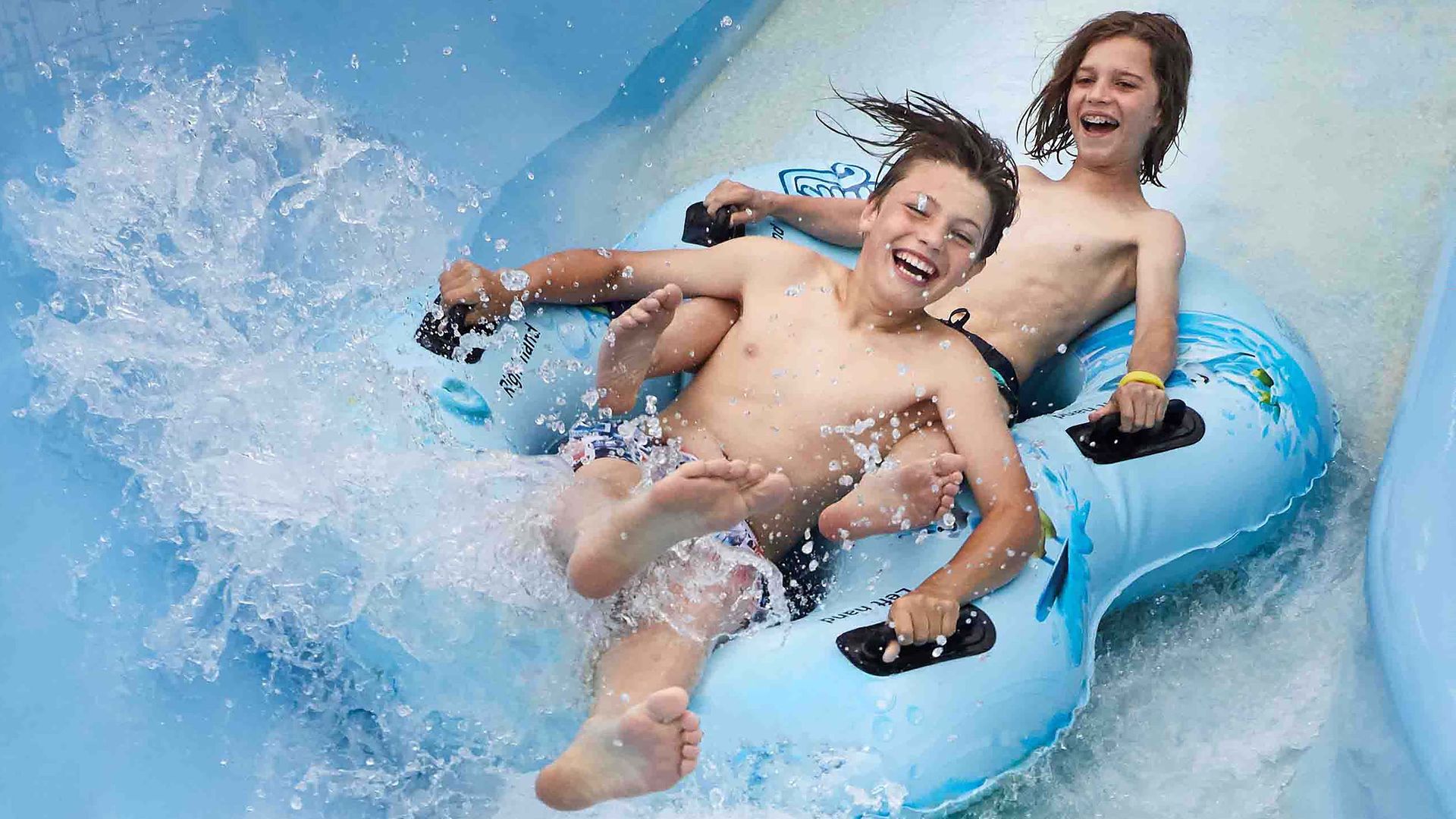 Zwei fröhliche Kinder rutschen auf einem aufblasbaren Reifen eine Wasserrutsche hinunter.
