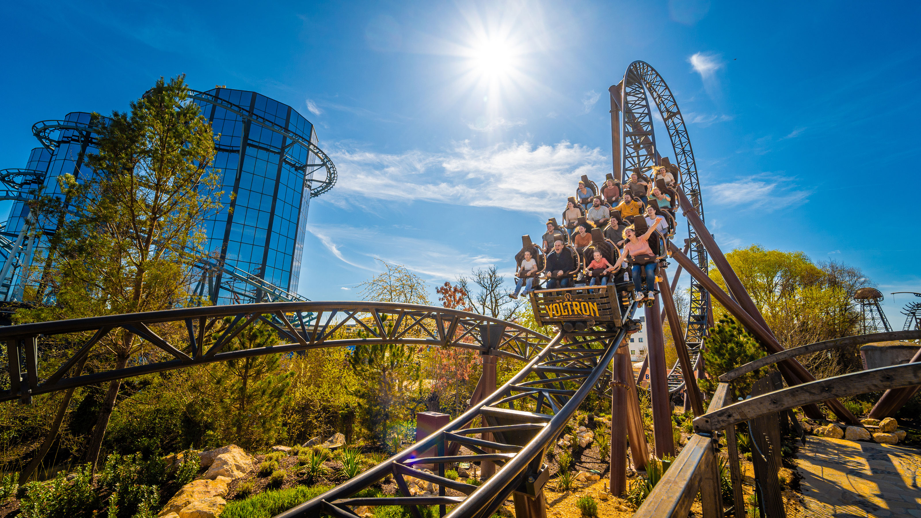 Roller coaster with passengers descending near a glass tower and trees under a sunny blue sky
