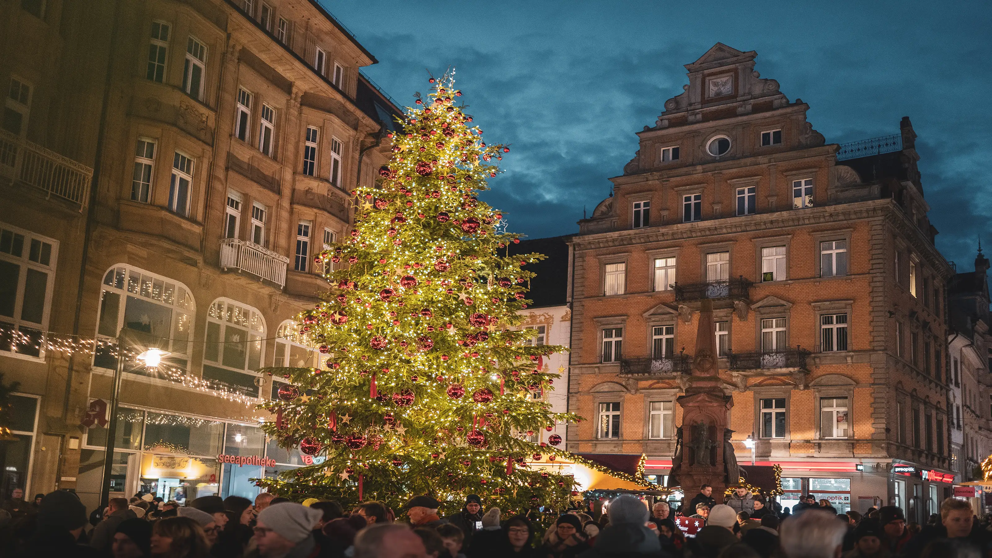 Sapin de Noël orné de guirlandes lumineuses et de boules, sur une place animée de la vieille ville, au crépuscule.