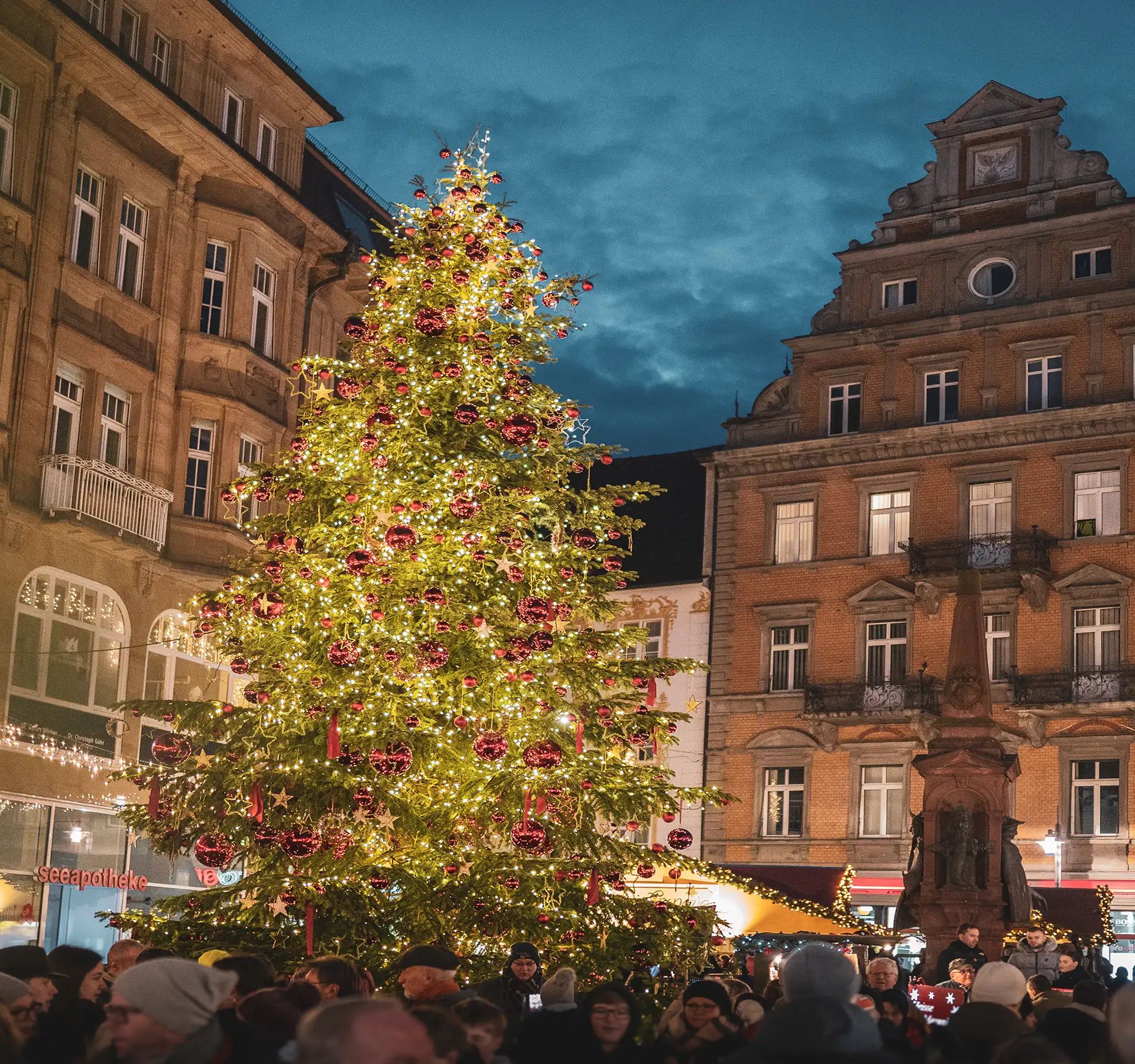 Sapin de Noël orné de guirlandes lumineuses et de boules, sur une place animée de la vieille ville, au crépuscule.
