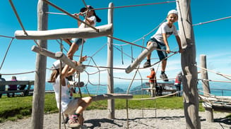 Three children play on a wooden climbing frame at a park with clear skies above and a scenic mountainous backdrop. the children are variously climbing and balancing, displaying expressions of enjoyment.