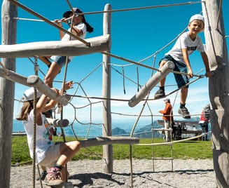 Drei Kinder spielen auf einem hölzernen Klettergerüst in einem Park mit klarem Himmel und einer malerischen Bergkulisse. Die Kinder klettern und balancieren auf verschiedene Weise und zeigen dabei einen Ausdruck von Vergnügen.