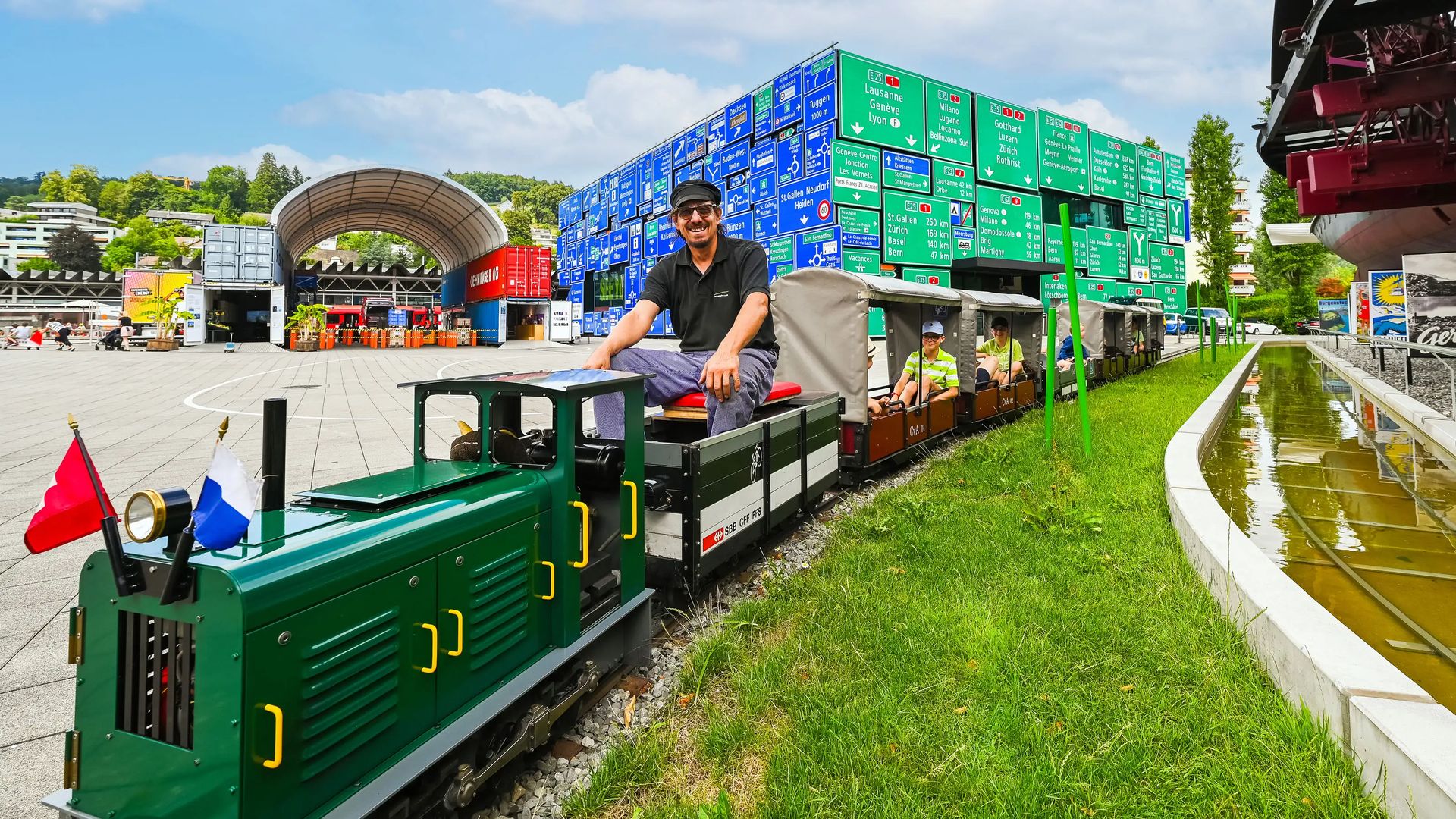 Chemin de fer miniature avec un train vert et plusieurs wagons ouverts, occupés par des enfants, le long des voies du Musée Suisse des Transports.