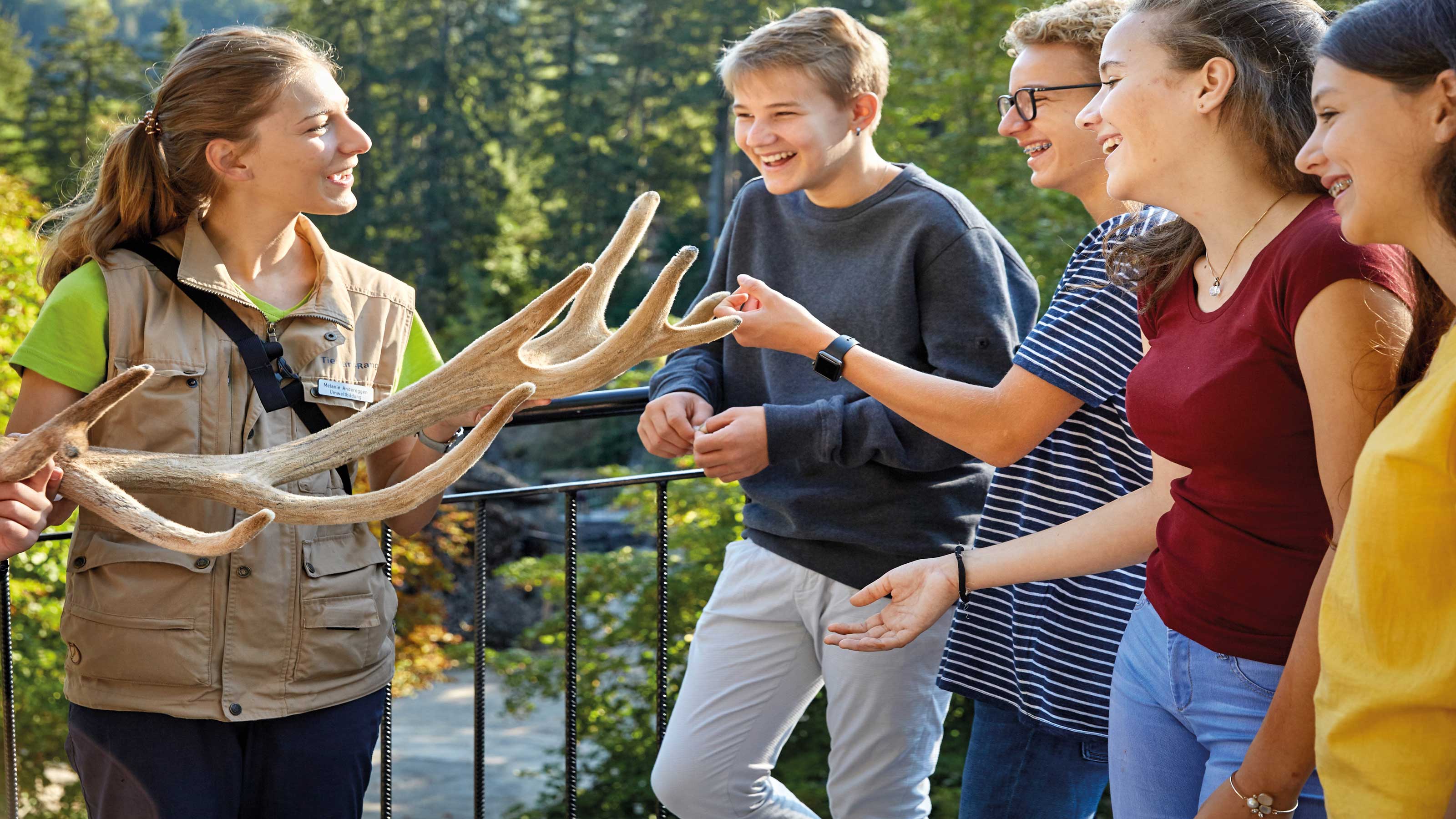 Eine junge Mitarbeiterin des Natur- und Tierpark Goldau hält ein grosses Geweih, während sie einer Gruppe Jugendlicher im Freien etwas erklärt.