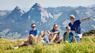 A family of five with a small dog, sitting on a grassy hillside, enjoys a sunny day with scenic mountains in the background.