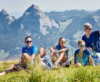 Eine fünfköpfige Familie mit einem kleinen Hund sitzt auf einer grasbewachsenen Anhöhe und genießt einen sonnigen Tag mit den malerischen Bergen im Hintergrund.