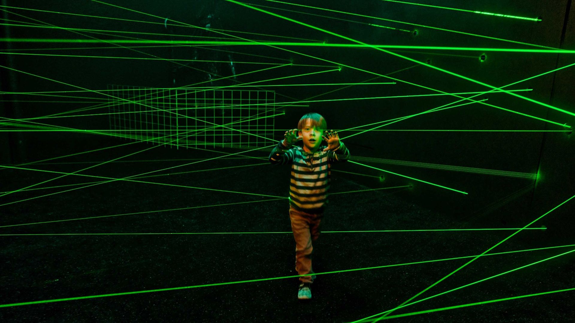 A young boy stands fascinated in a dark room with lots of green lasers.