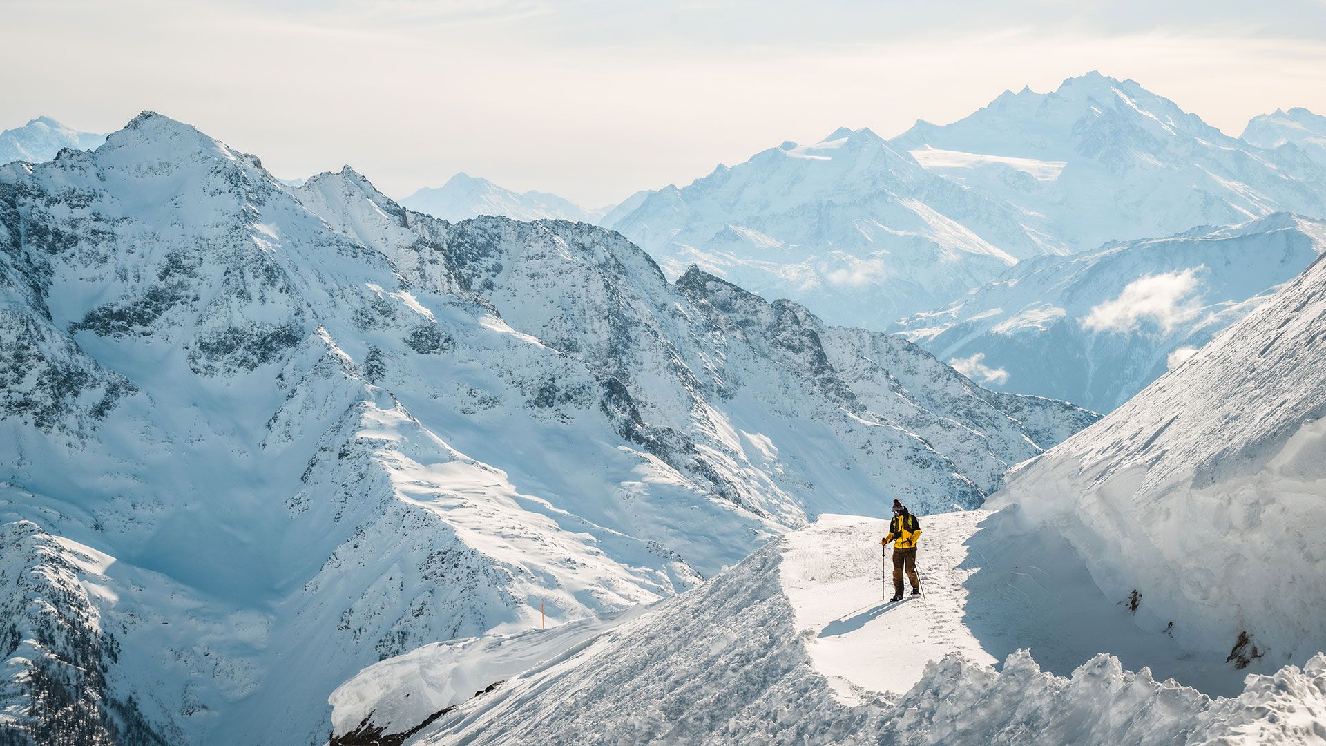 A person stands triumphantly atop a snow-covered mountain, surrounded by majestic peaks under a clear blue sky.