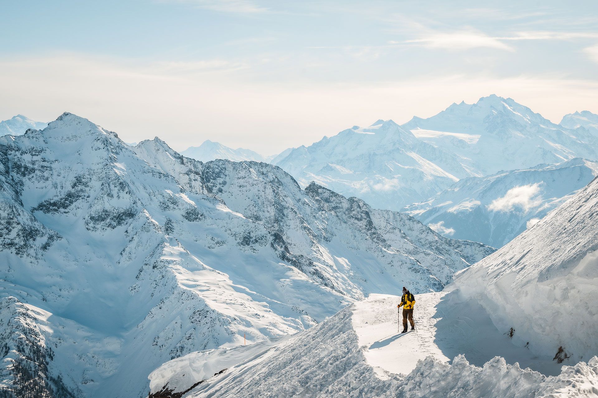 Une personne se tient triomphalement au sommet d'une montagne enneigée, entourée de pics majestueux sous un ciel bleu limpide.
