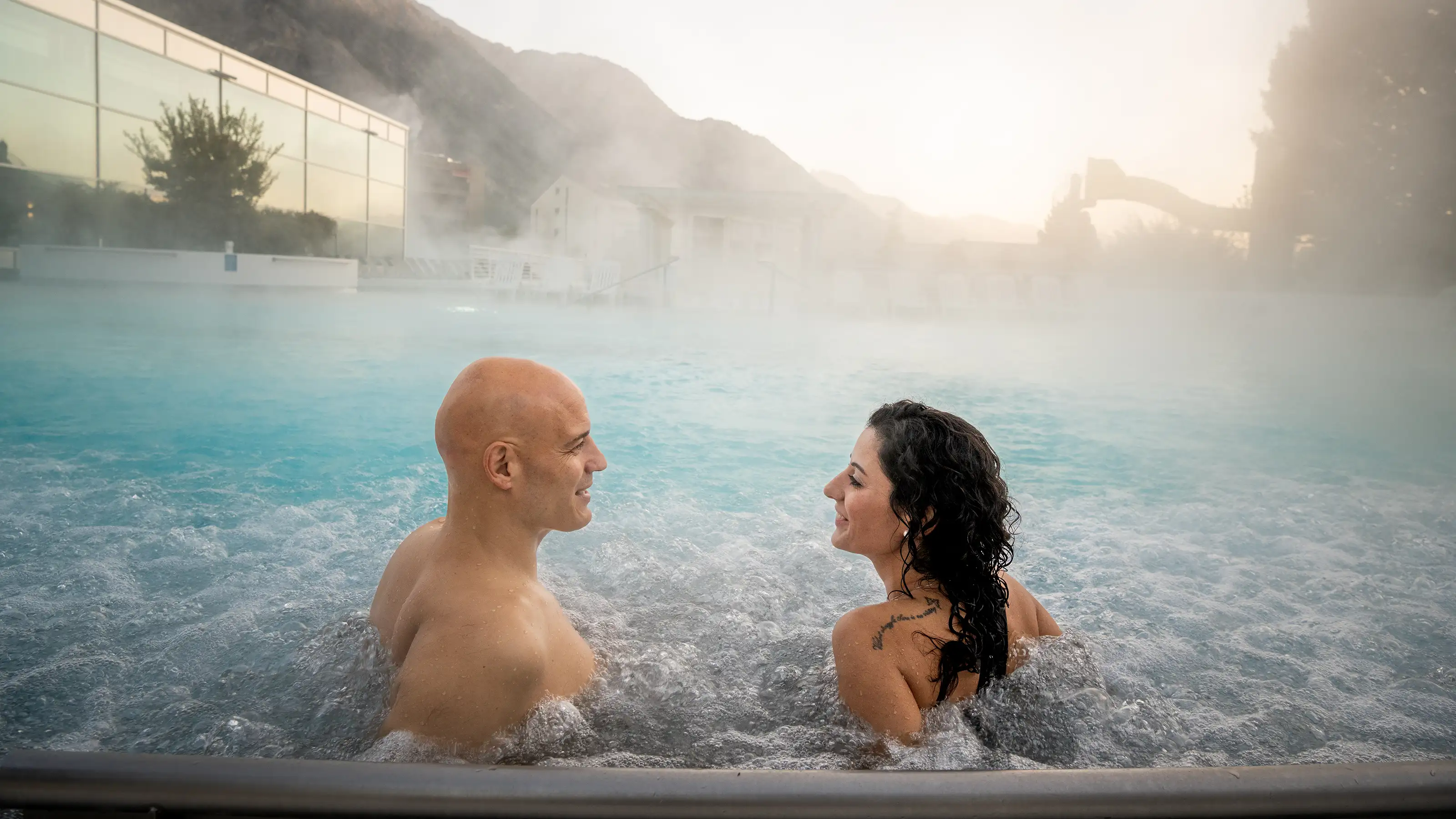 Zwei Personen sitzen im Aussenpool der Bains de Saillon mit dampfendem Wasser und Berglandschaft im Hintergrund.
