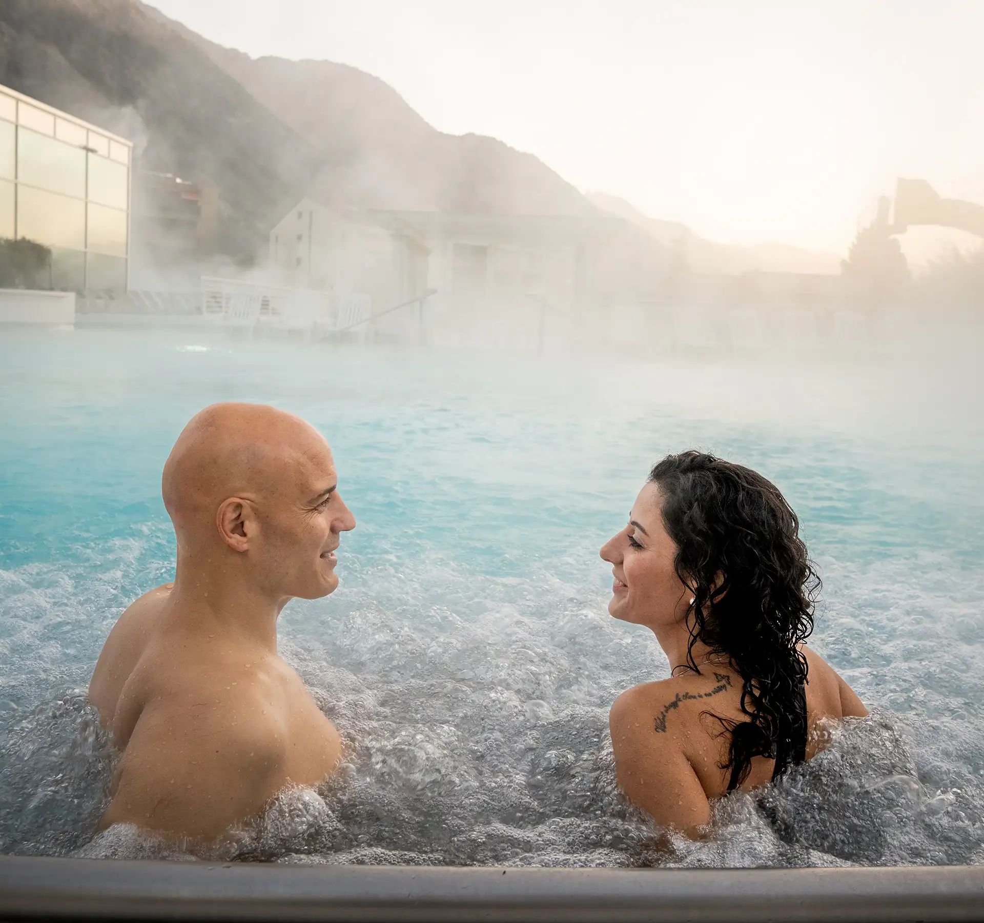 Two people sitting in the outdoor pool at Bains de Saillon with steaming water and mountains in the background.