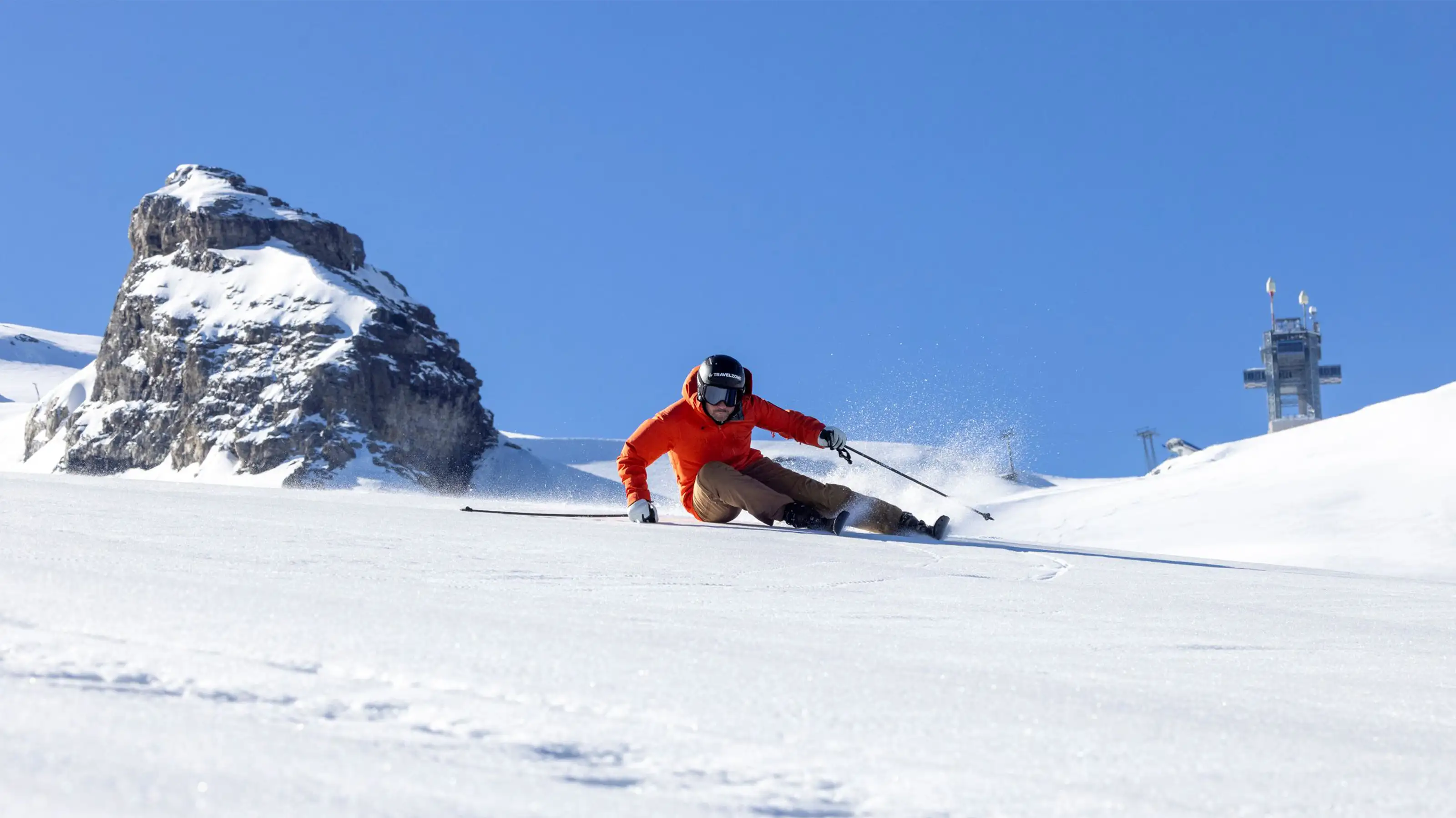 Skifahrer in roter Jacke fährt auf verschneiter Piste vor Felsen und blauem Himmel.