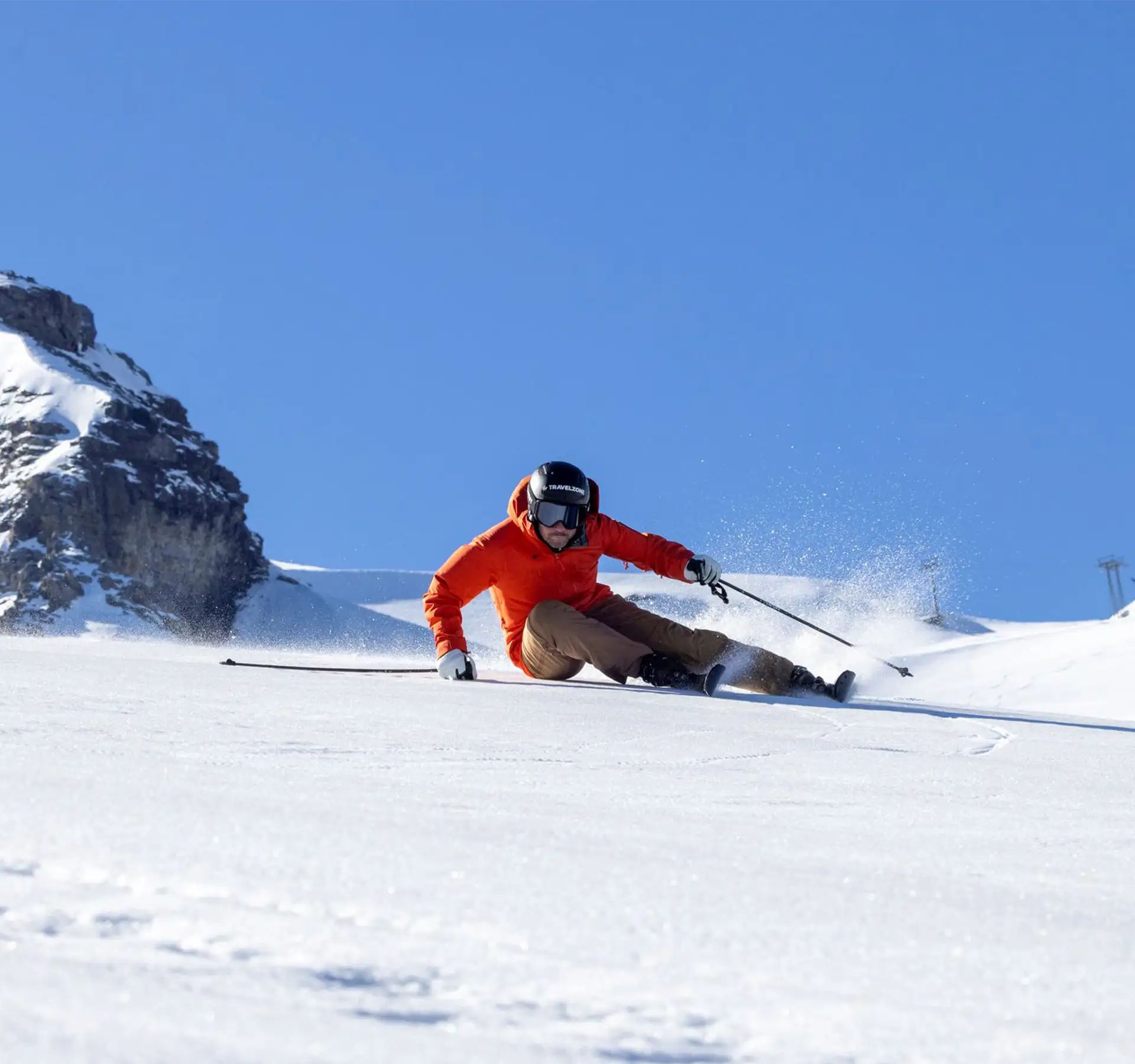 Skifahrer in roter Jacke fährt auf verschneiter Piste vor Felsen und blauem Himmel.