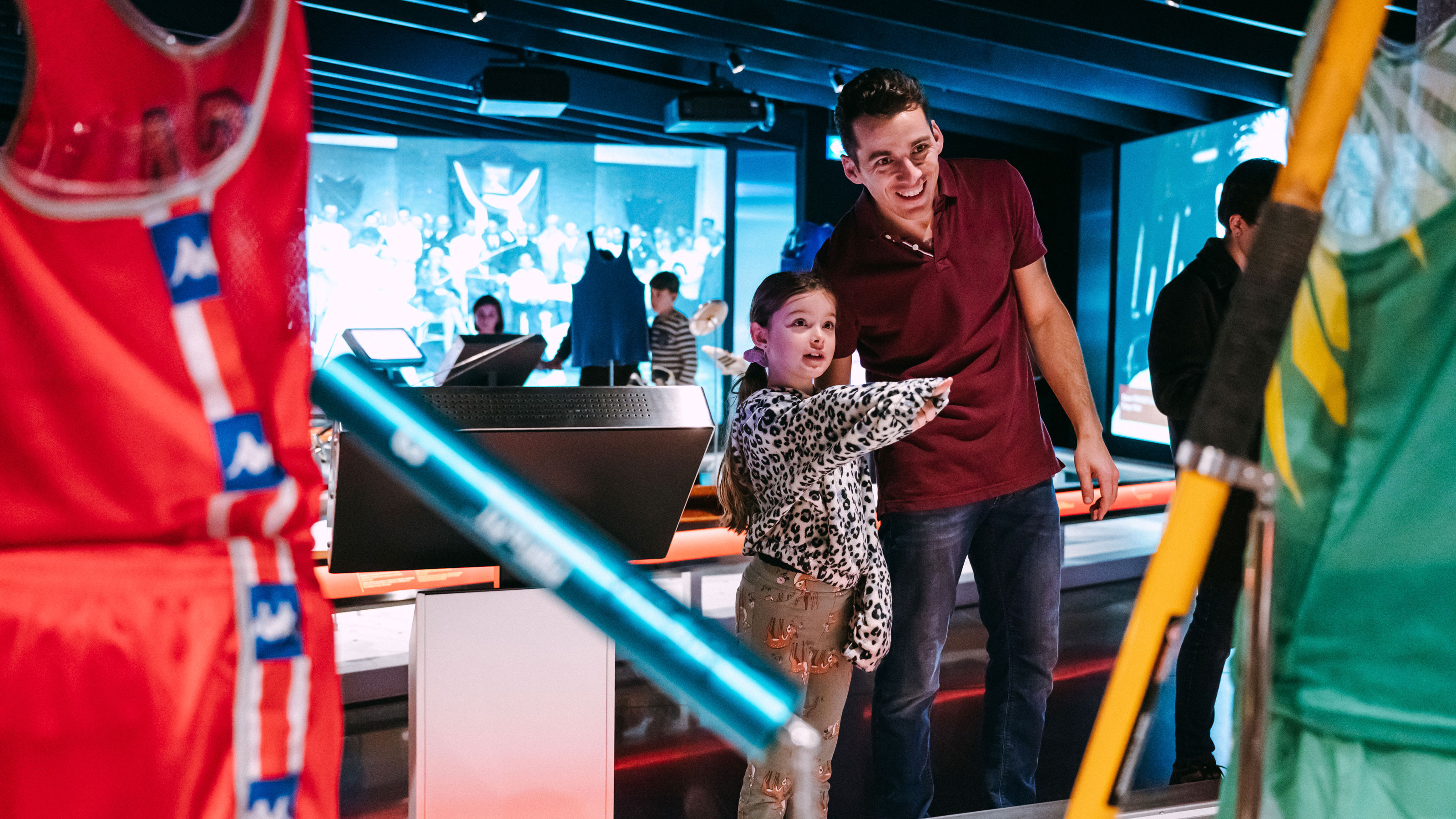A man stands smiling next to a young girl who is excitedly pointing at something outside the picture, surrounded by colourful displays and screens in a lively, interactive exhibition.