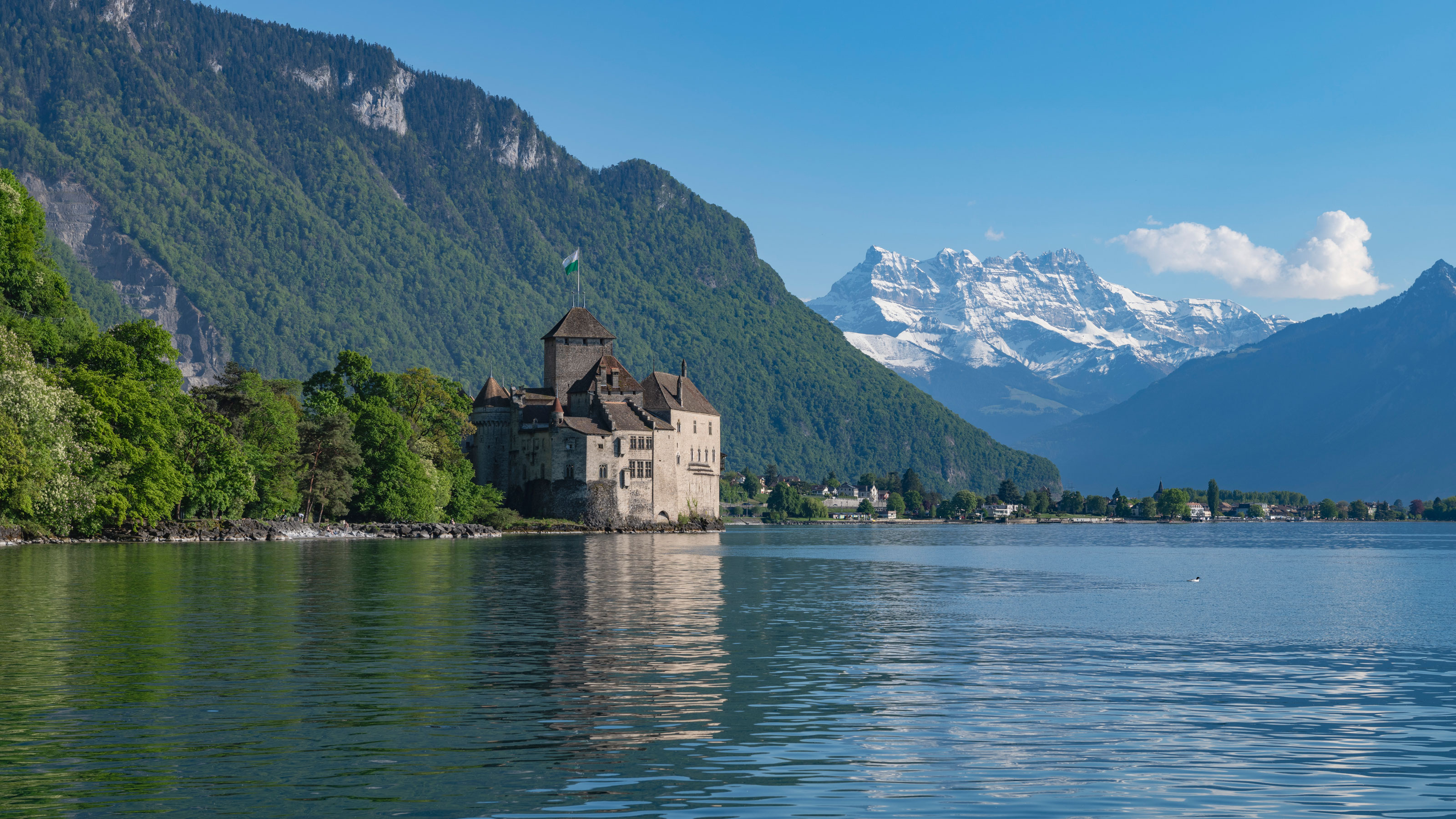 Schloss am Ufer eines Sees, umgeben von bewaldeten Bergen und schneebedeckten Gipfeln im Hintergrund