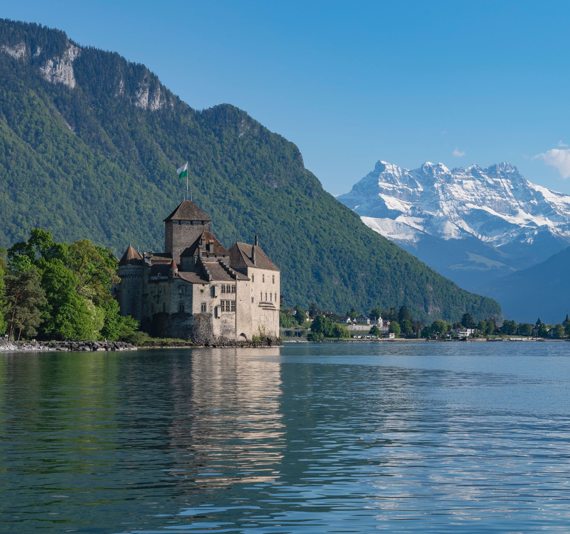 Schloss am Ufer eines Sees, umgeben von bewaldeten Bergen und schneebedeckten Gipfeln im Hintergrund