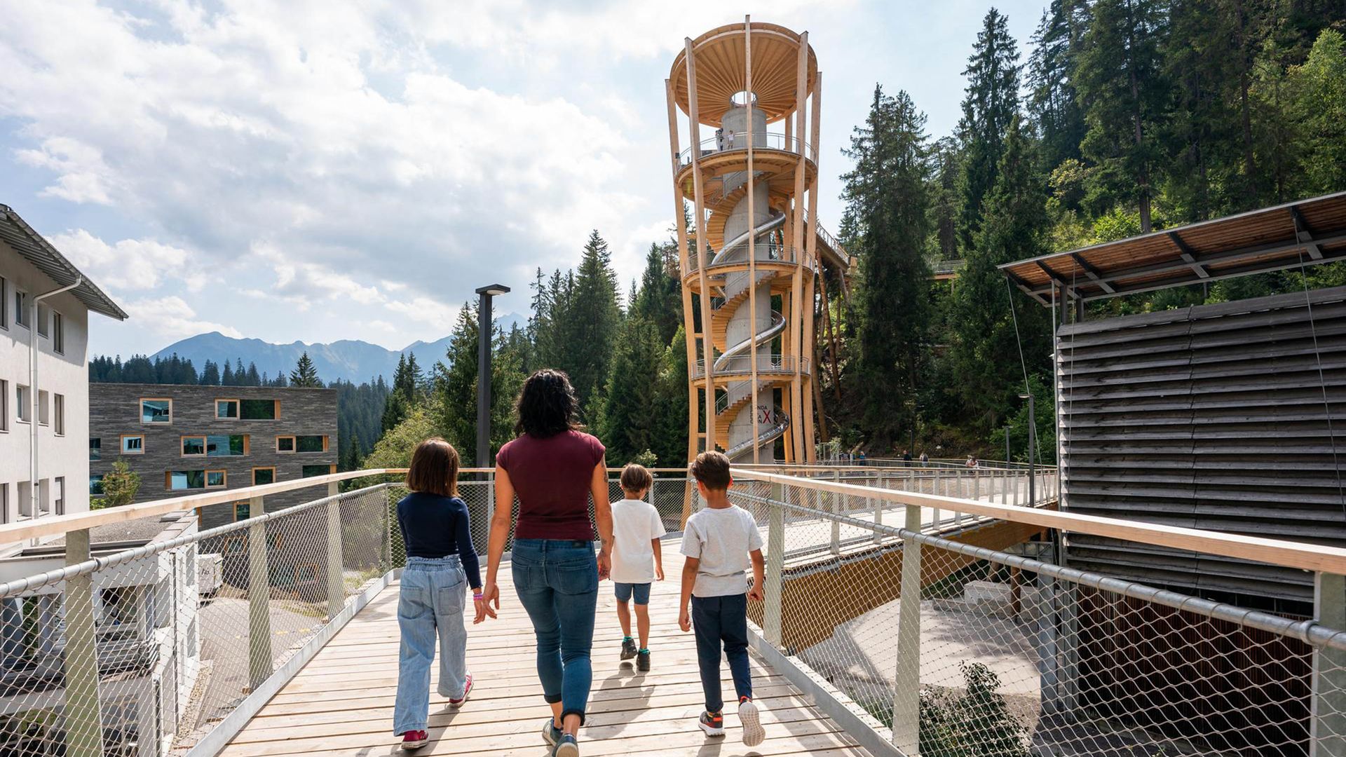 A family walks along the wooden walkway of the Laax treetop path towards a viewing tower.