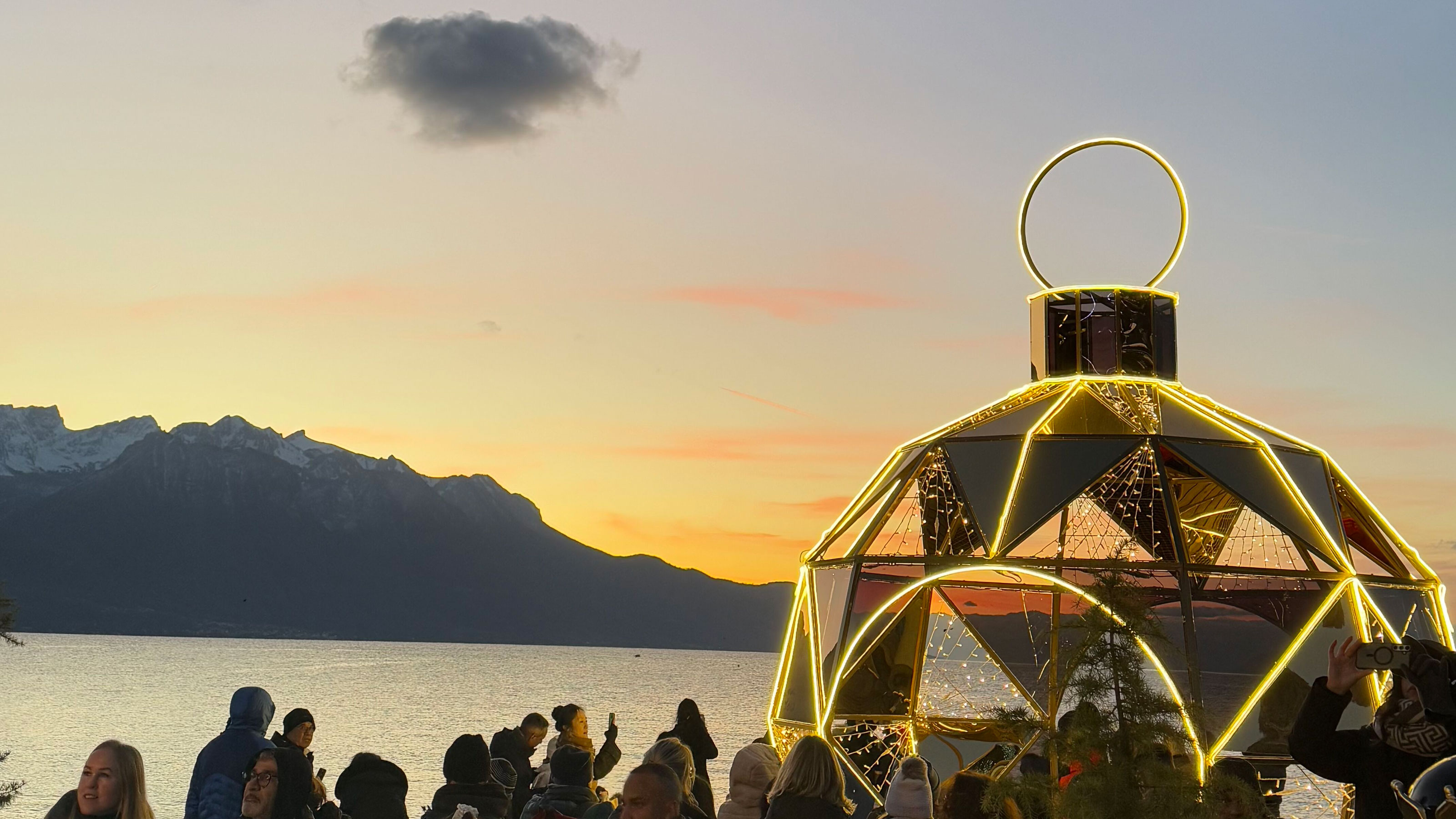 Coucher de soleil au bord du lac avec les montagnes en arrière-plan, des gens devant une grande décoration de Noël
