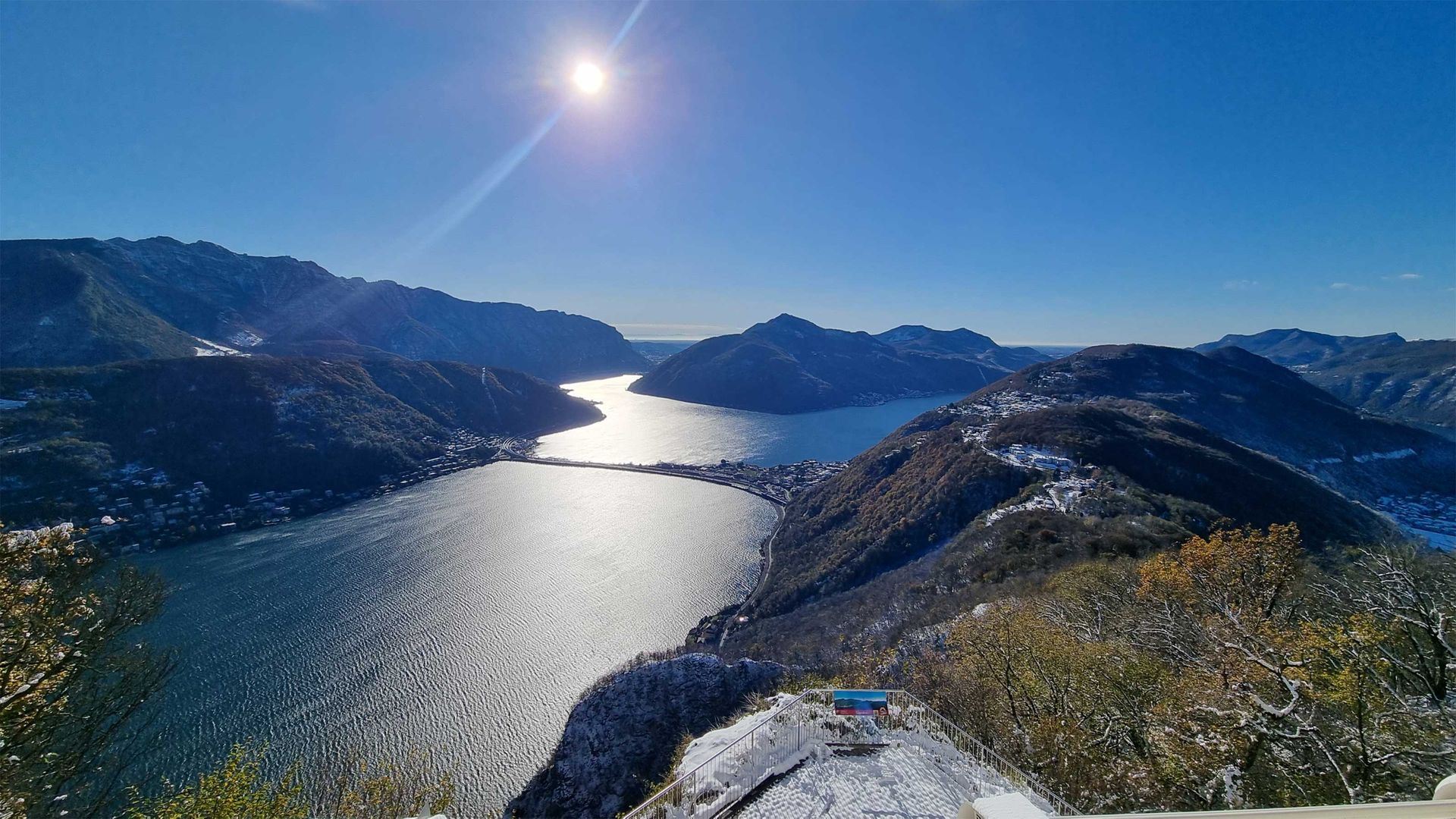 View of a sunlit landscape with a lake surrounded by snow-covered mountains and autumnal forests.