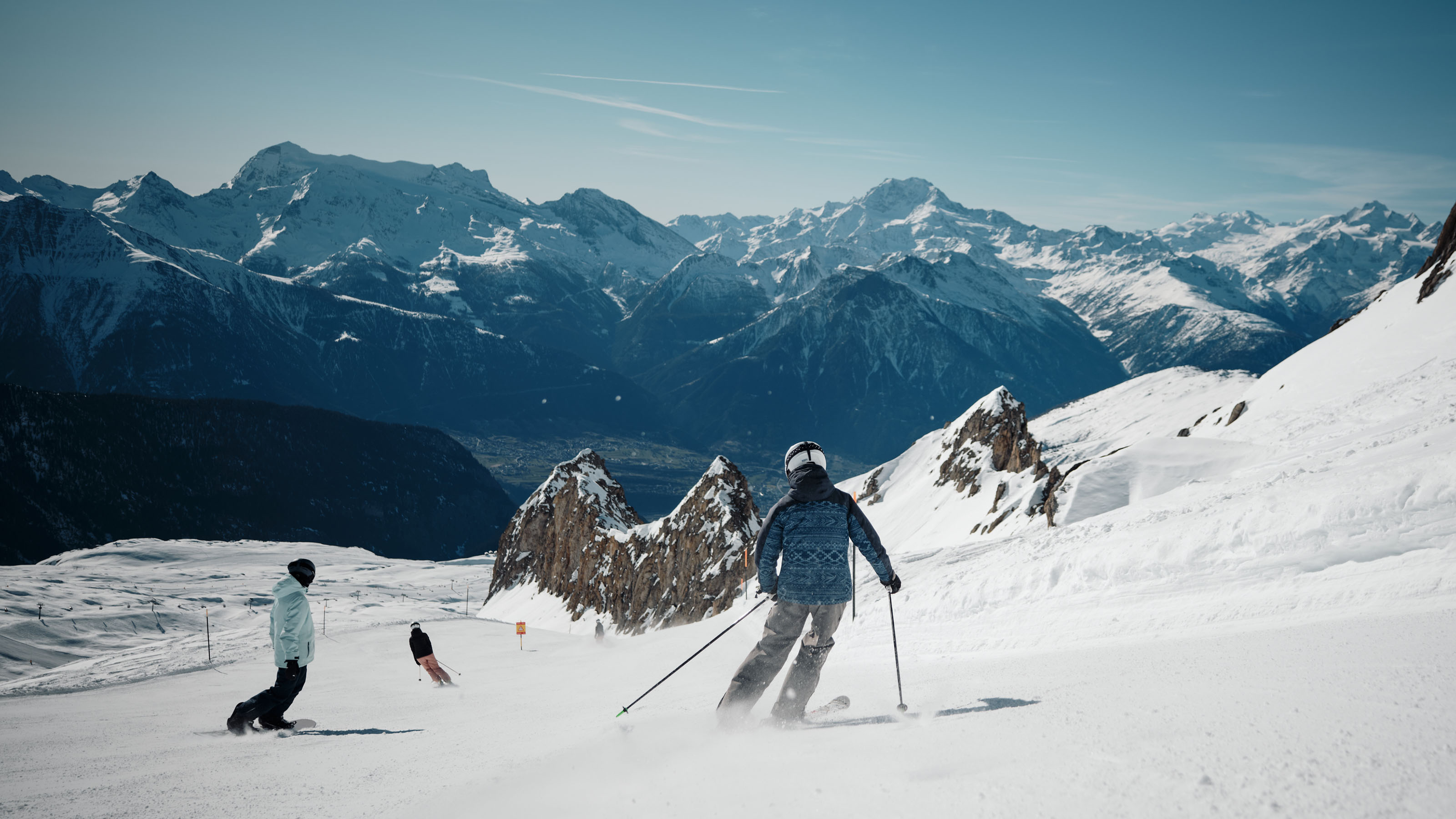 Mehrere Skifahrer fahren auf einer schneebedeckten Piste im Skigebiet Blatten Belalp mit Bergpanorama im Hintergrund.