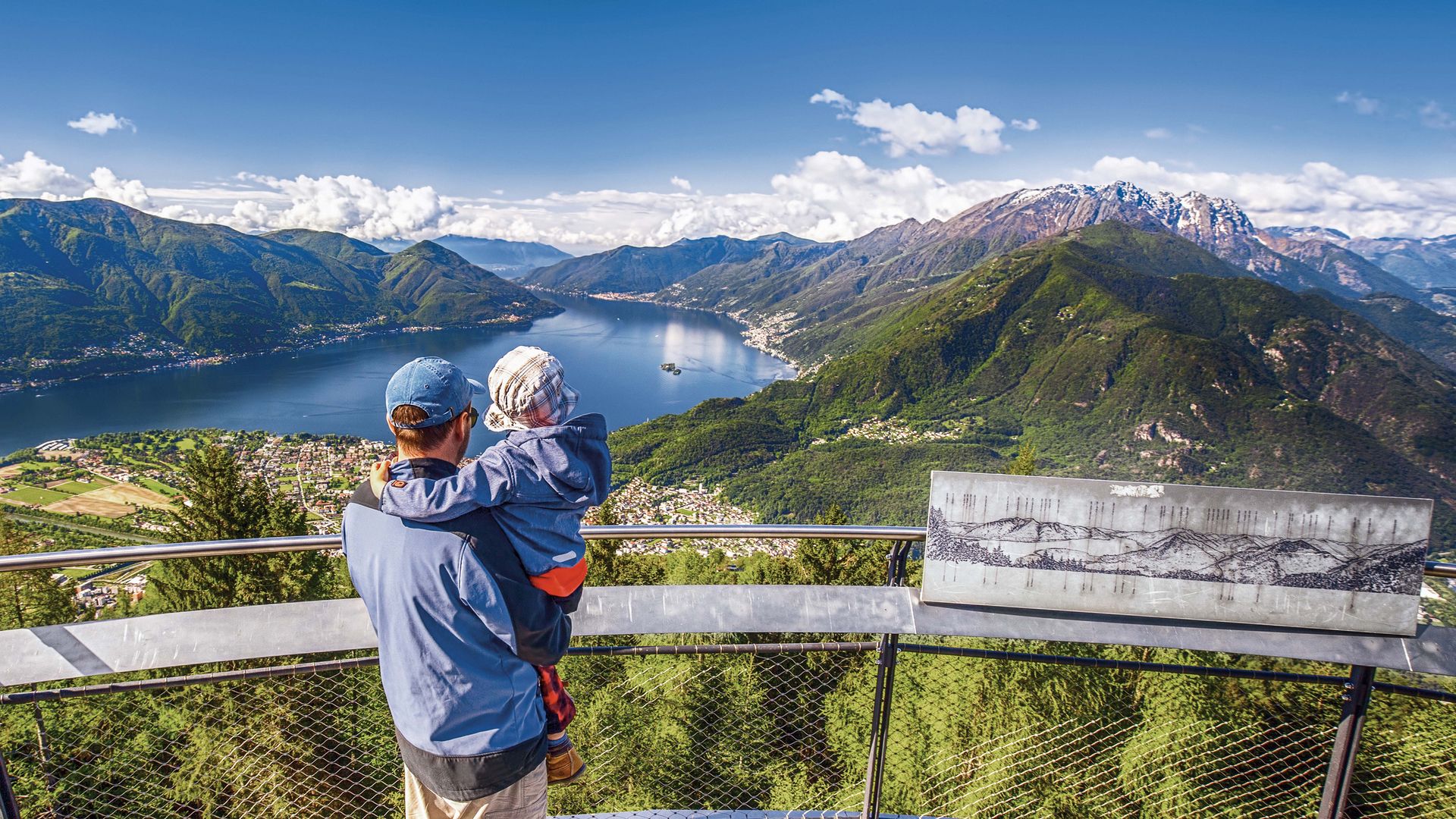 Ein Mann hält sein Kind im Arm und sie geniessen die Aussicht auf den Lago Maggiore und die umliegenden Täler.