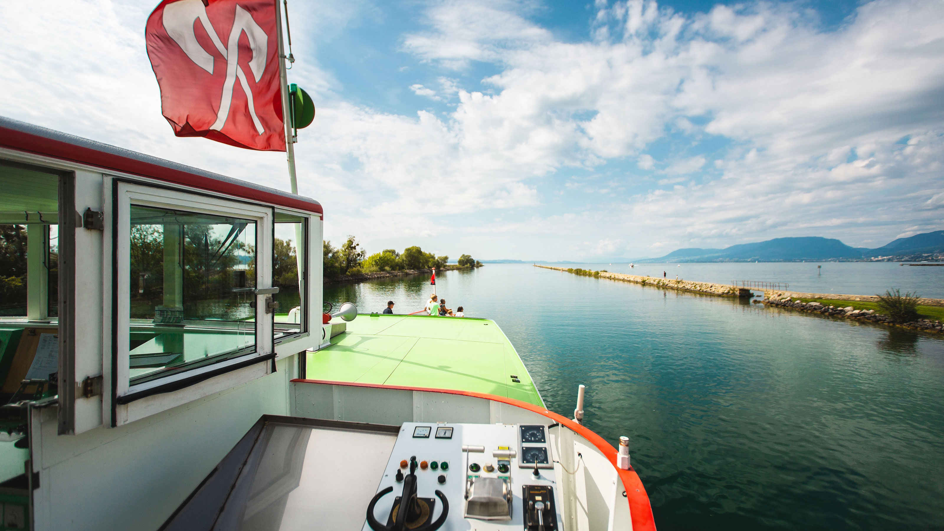 Das Bild zeigt das Deck eines Bootes mit einer roten Flagge, das auf einem ruhigen Gewässer mit grünen Ufern und Bergen im Hintergrund fährt.
