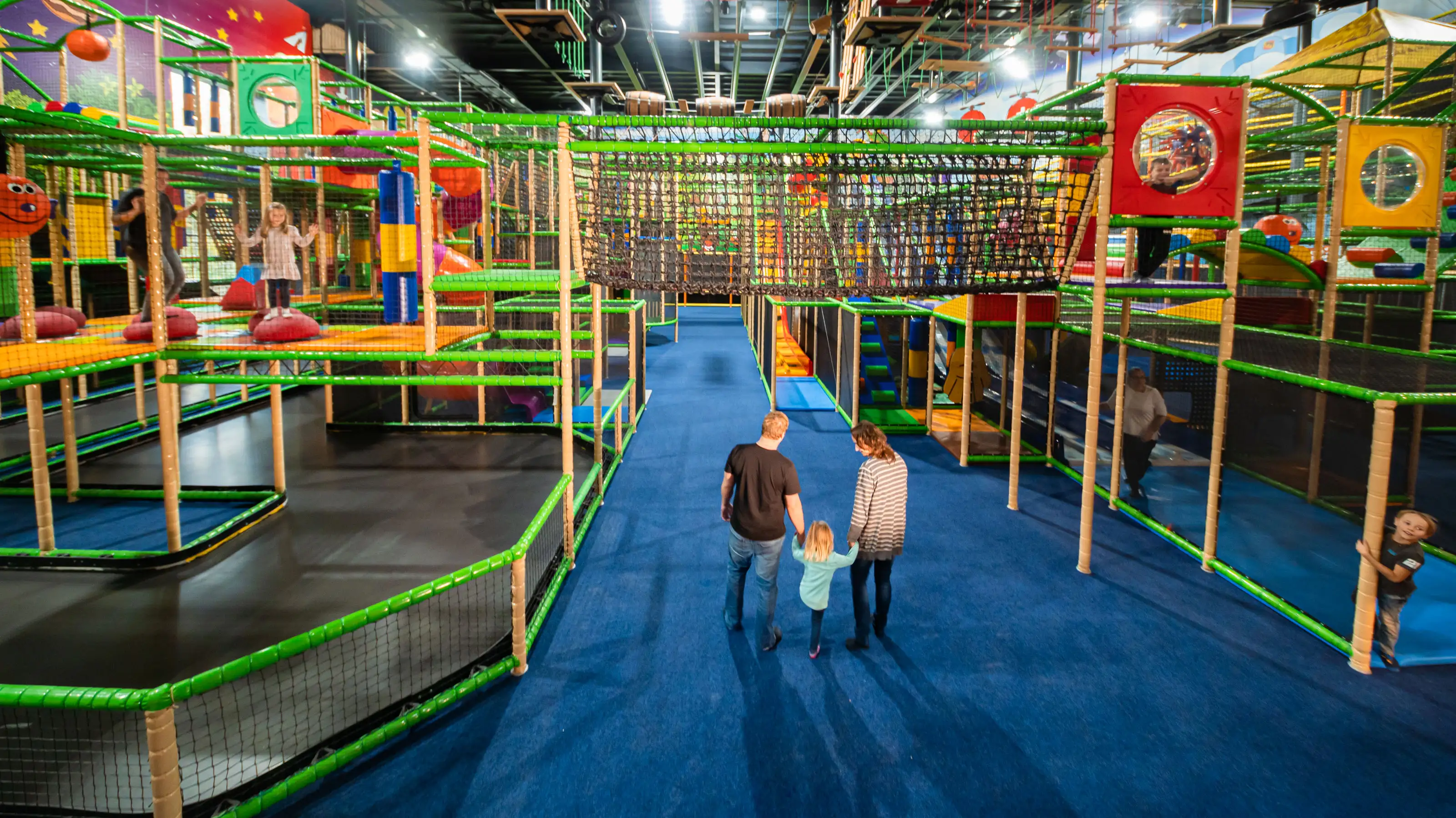 Vue intérieure du Kiddy Dome avec des structures d'escalade colorées et trois personnes marchant sur un sol bleu vers elles