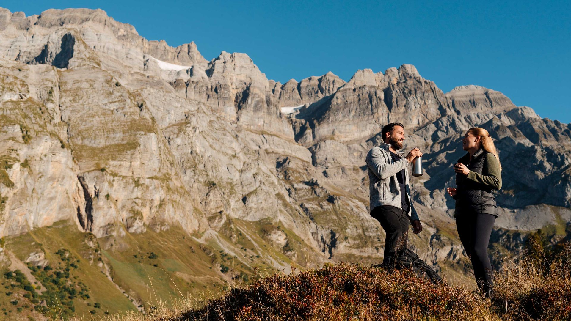Zwei Wanderer machen eine Pause, trinken etwas und unterhalten sich auf einem Bergpfad mit majestätischen Felsgipfeln im Hintergrund.