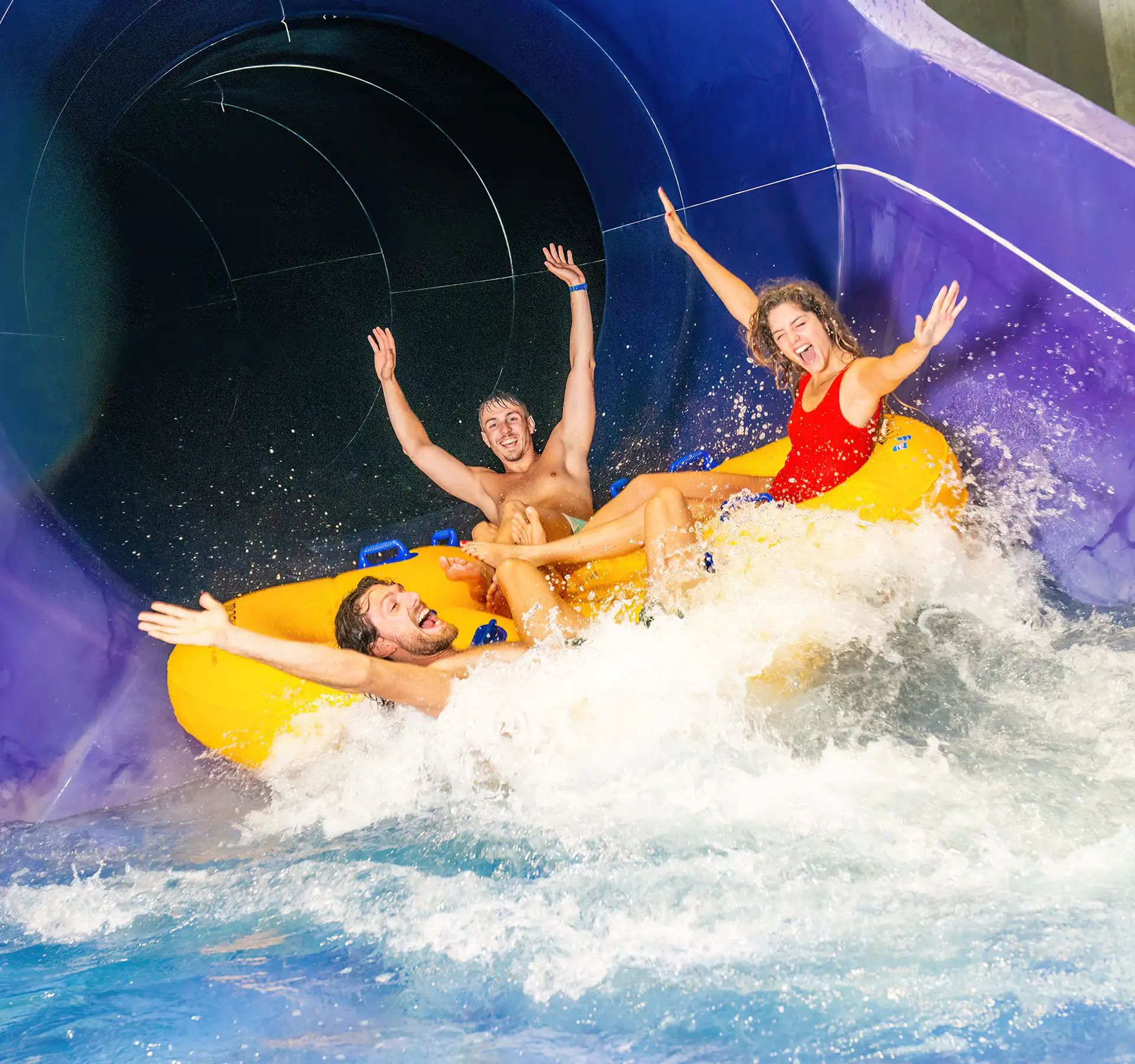 Three people on a yellow inflatable raft exiting a purple water slide into a pool