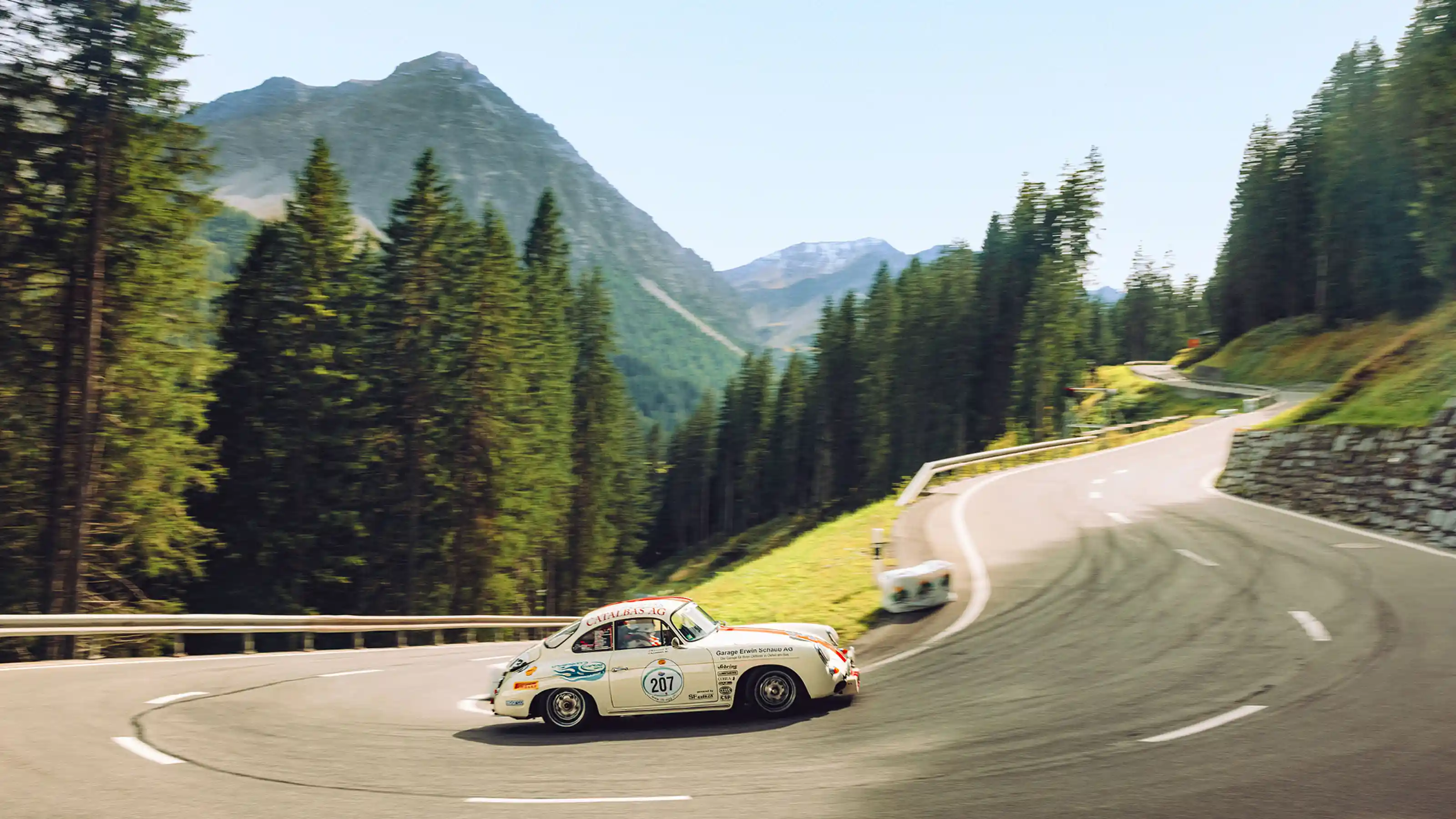 Auto sportiva classica su strada di montagna tortuosa con montagne boschive sullo sfondo.