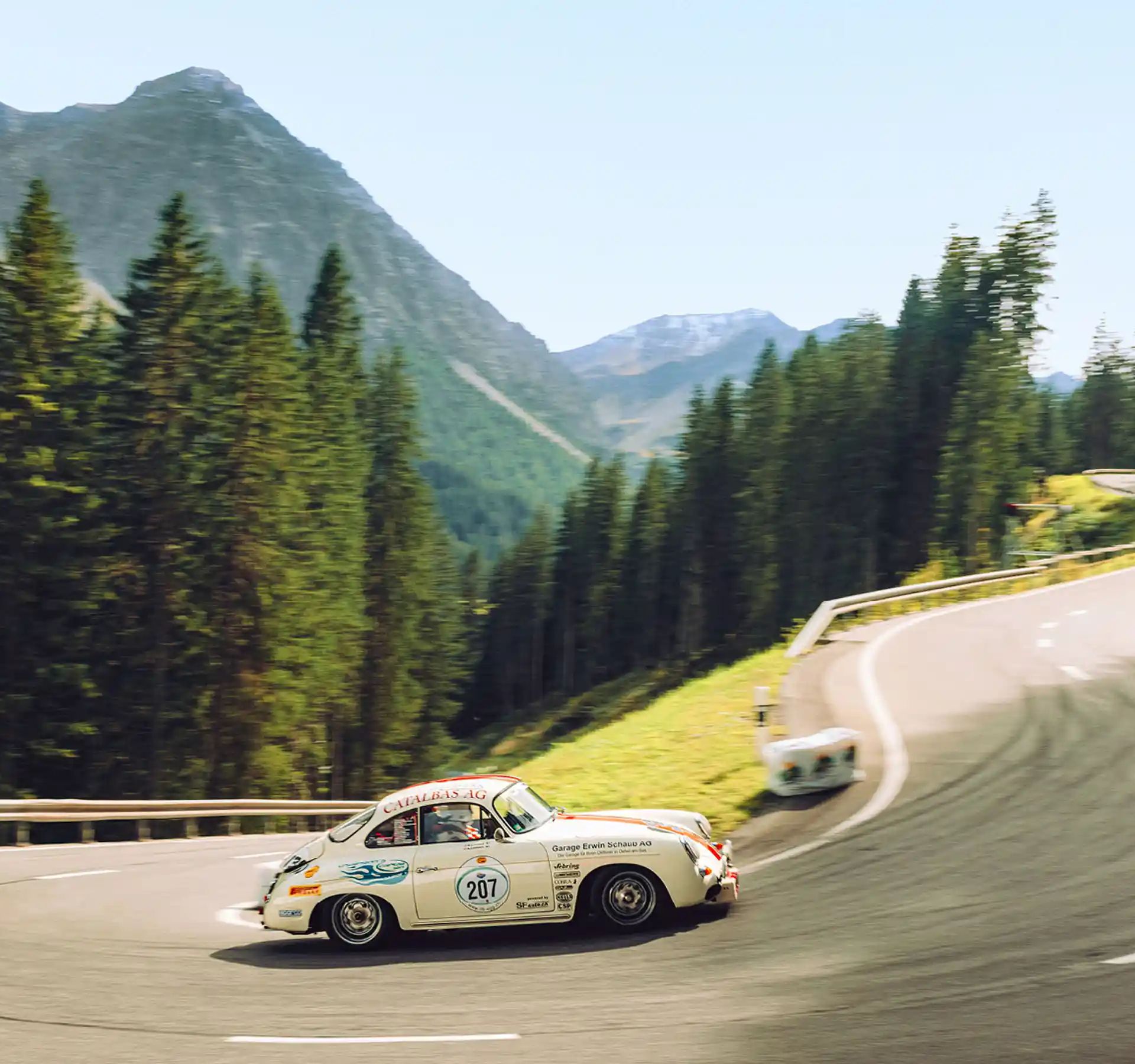 Classic sports car on winding mountain road with forested mountains in the background.