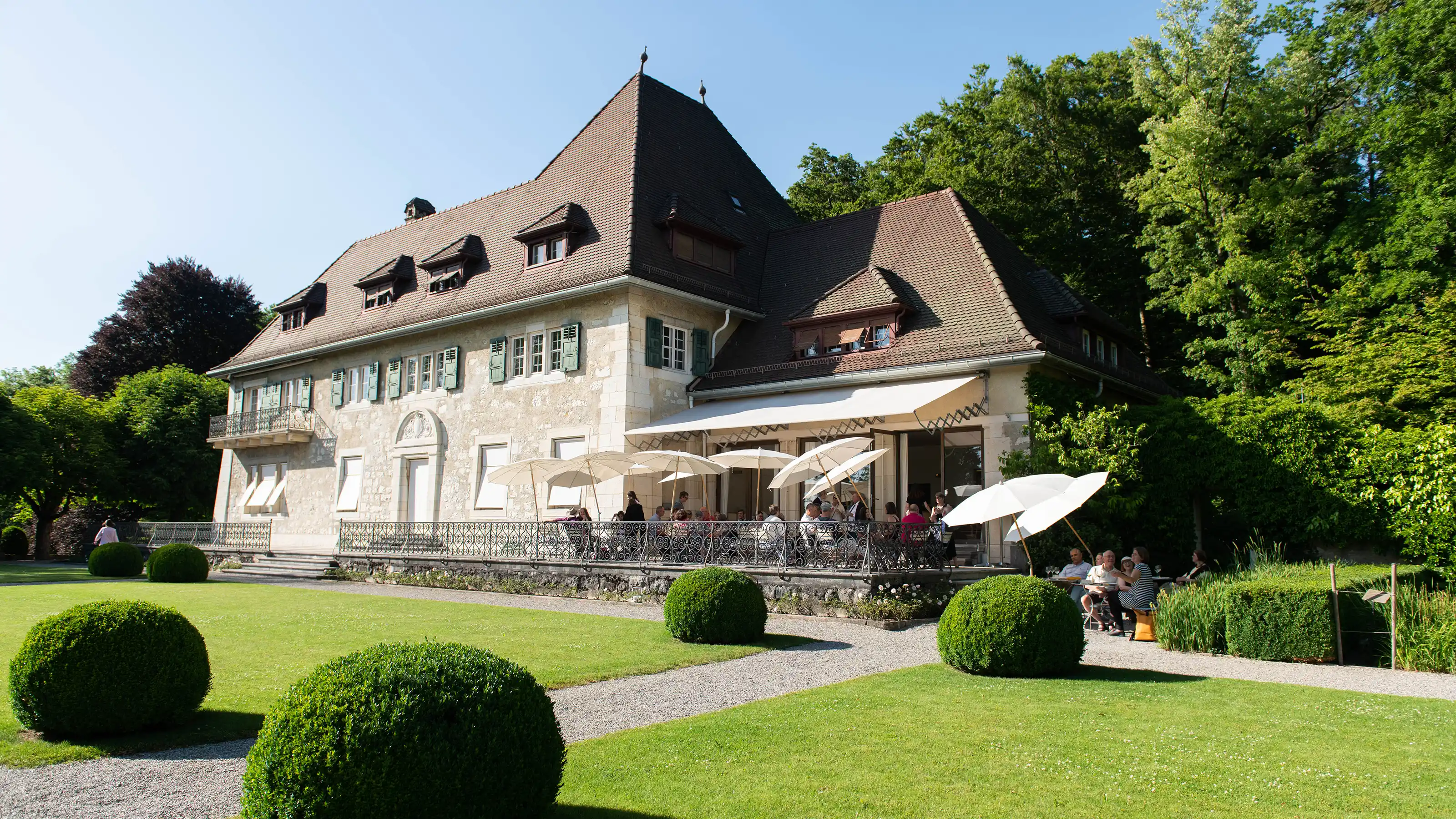 Grosses historisches Gebäude mit steilem Dach und Terrasse mit Sonnenschirmen vor gepflegtem Rasen und kugelförmigen Hecken.