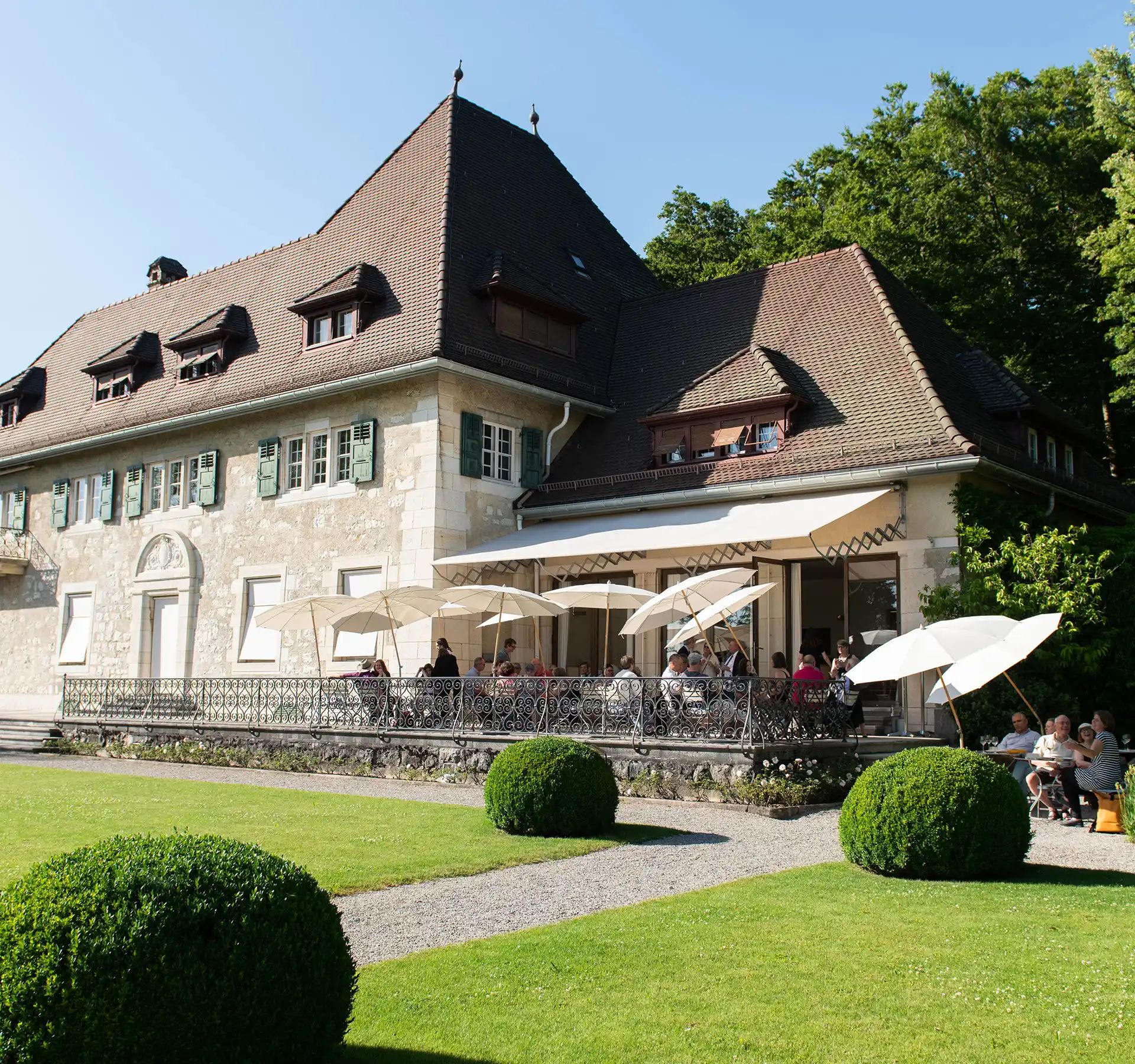 Grosses historisches Gebäude mit steilem Dach und Terrasse mit Sonnenschirmen vor gepflegtem Rasen und kugelförmigen Hecken.