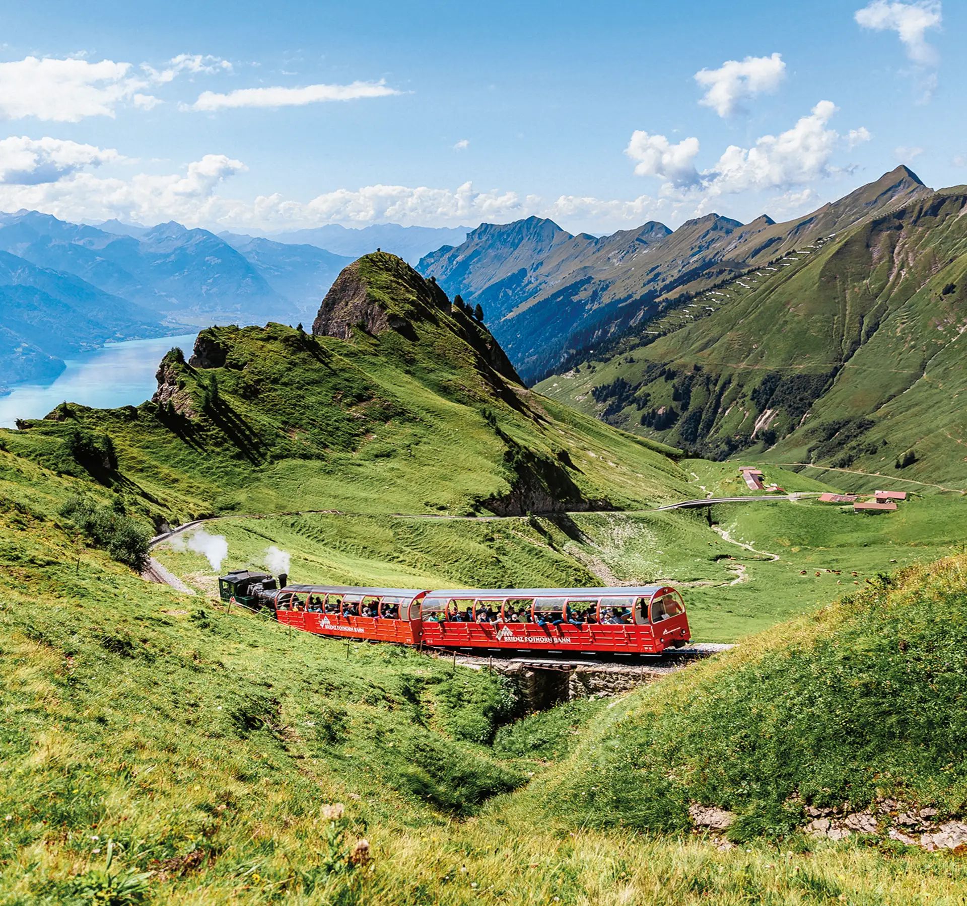 The red cable car of the Brienzer Rothornbahn crosses a bridge through green Alpine countryside with mountains in the background.