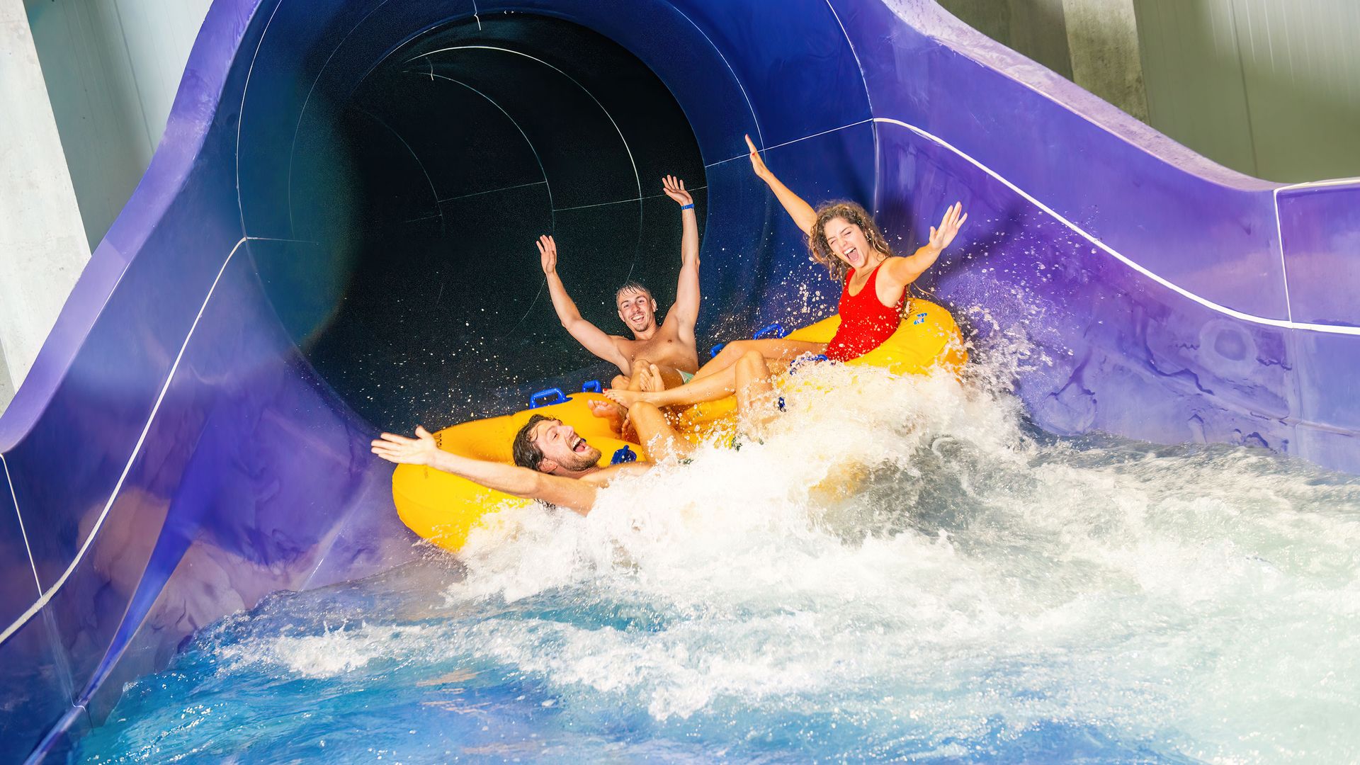 Tre persone su un gommone giallo che esce da uno scivolo d'acqua viola in una piscina
