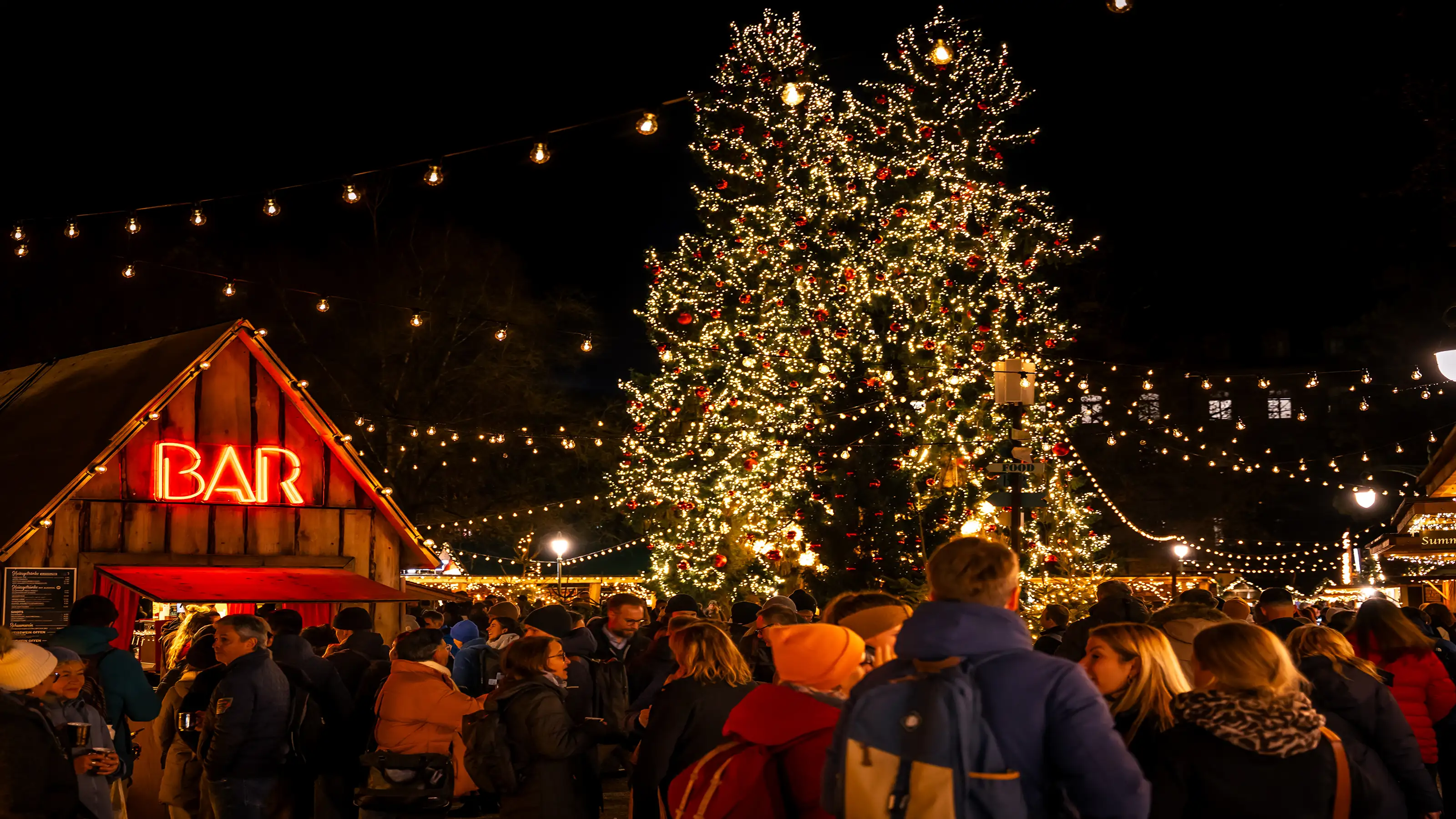  Mercatino di Natale con un grande albero di Natale addobbato, una capanna-bar illuminata e tanta gente di notte.