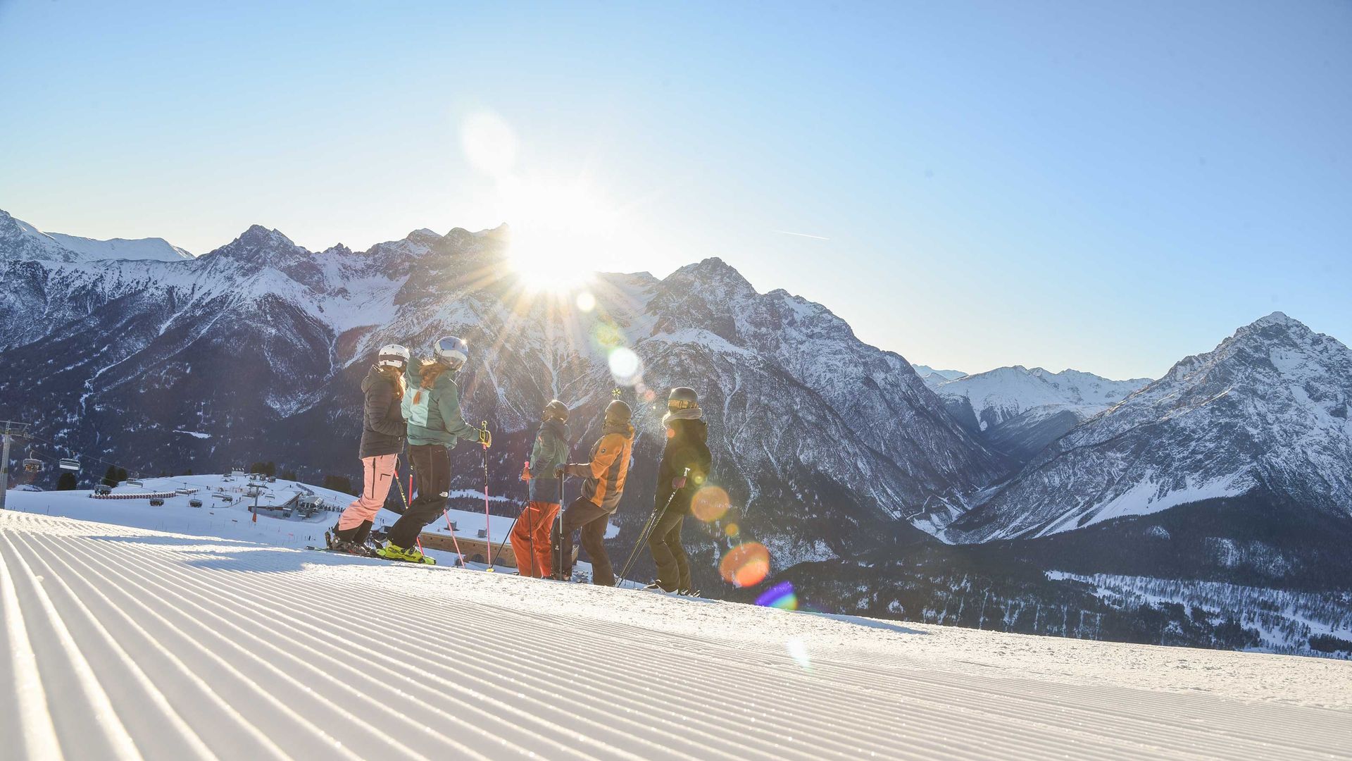 Des skieurs en vêtements d'hiver colorés se tiennent sur une piste fraîchement damée dans la station de ski Scuol Motta Naluns, devant des montagnes enneigées sous un soleil éclatant.