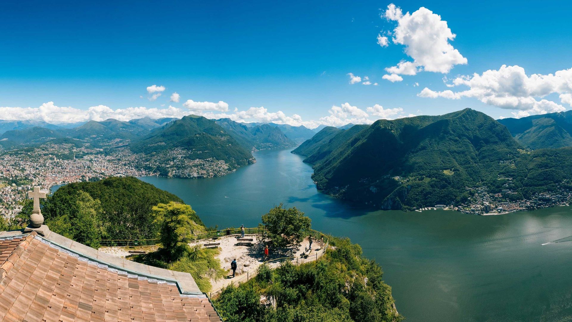 Panoramablick vom Monte San Salvatore auf den von Bergen umgebenen Luganersee mit Besuchern, die die Landschaft von einem hohen Aussichtspunkt aus genießen.