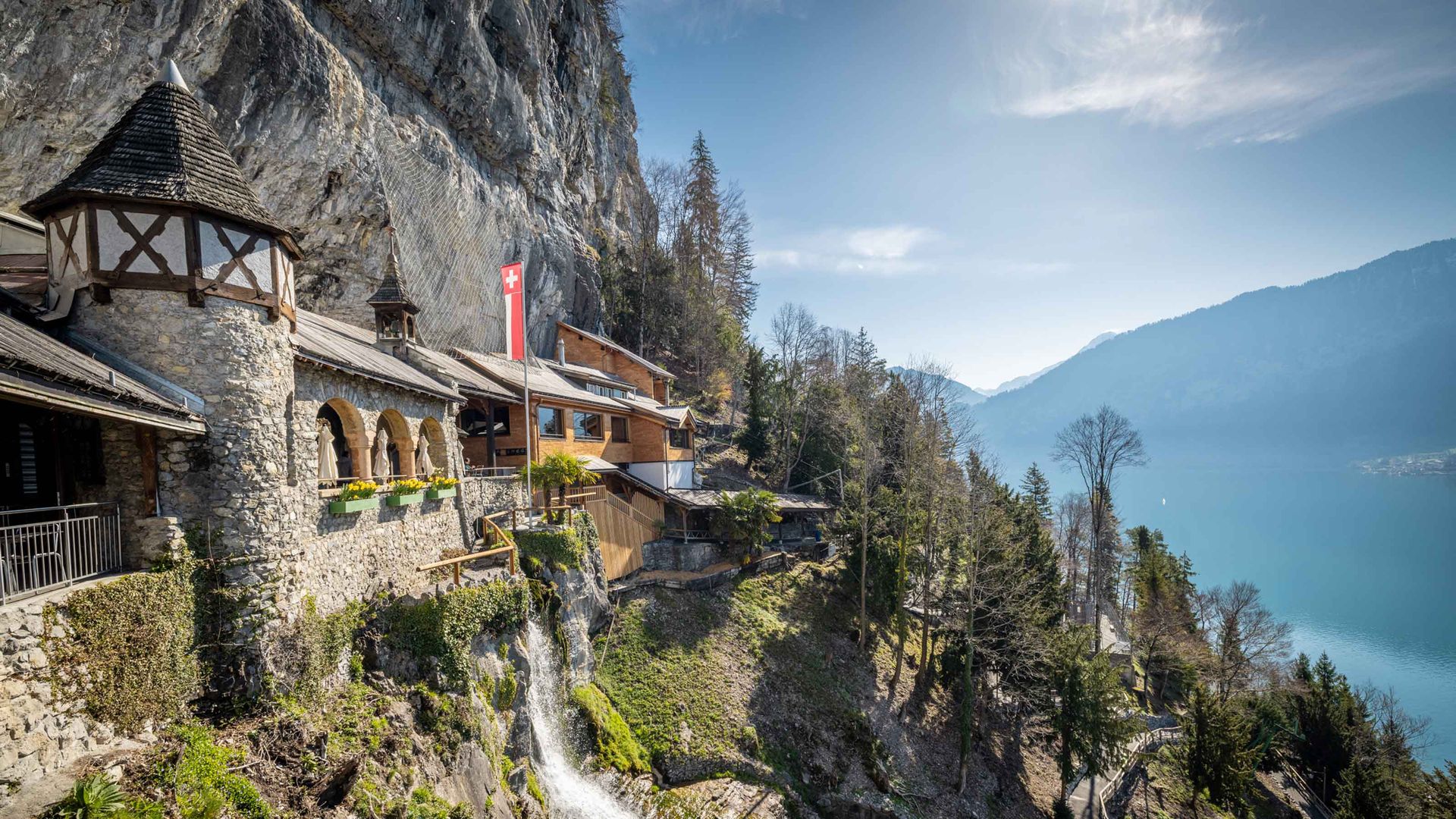 Aussenansicht der Beatushöhlen. An einer imposanten Felswand steht ein aus Stein gemauerter Turm neben einem neuen Gebäude aus Holz. Unterhalb fliesst ein kleiner Wasserfall die Felswand hinunter.
