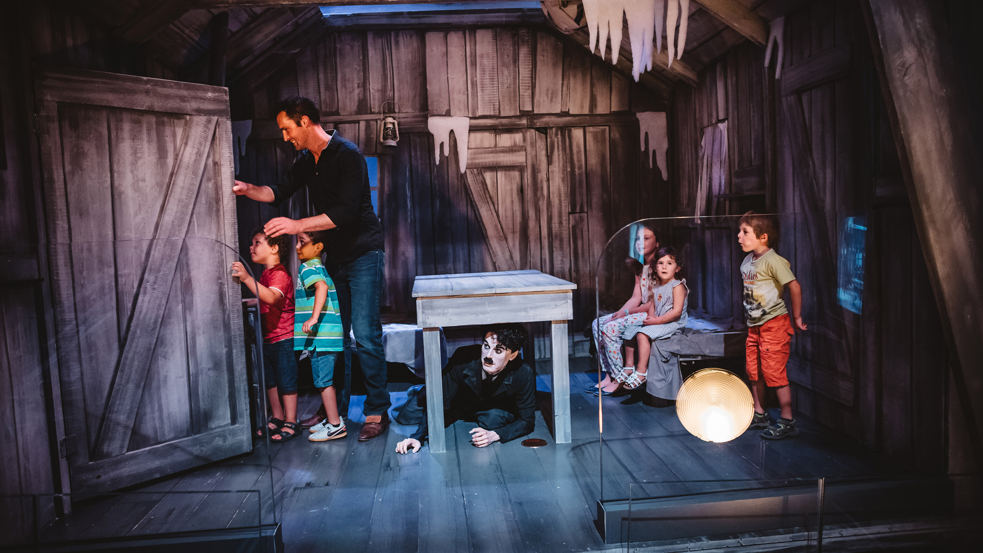 Ein Vater öffnet eine Tür auf einem Dachstock. Zwei Jungen schauen gespannt was sich dahinter versteckt. Drei weiter Kinder beobachten das Geschehen von weiter hinten und Charlie lugt unter einem Tisch hervor.