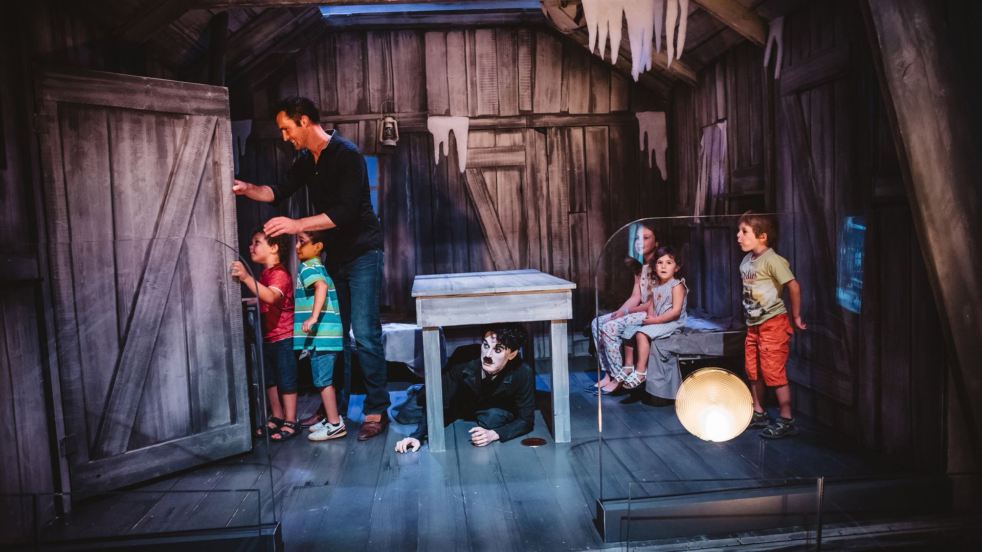 Ein Vater öffnet eine Tür auf einem Dachstock. Zwei Jungen schauen gespannt was sich dahinter versteckt. Drei weiter Kinder beobachten das Geschehen von weiter hinten und Charlie lugt unter einem Tisch hervor.