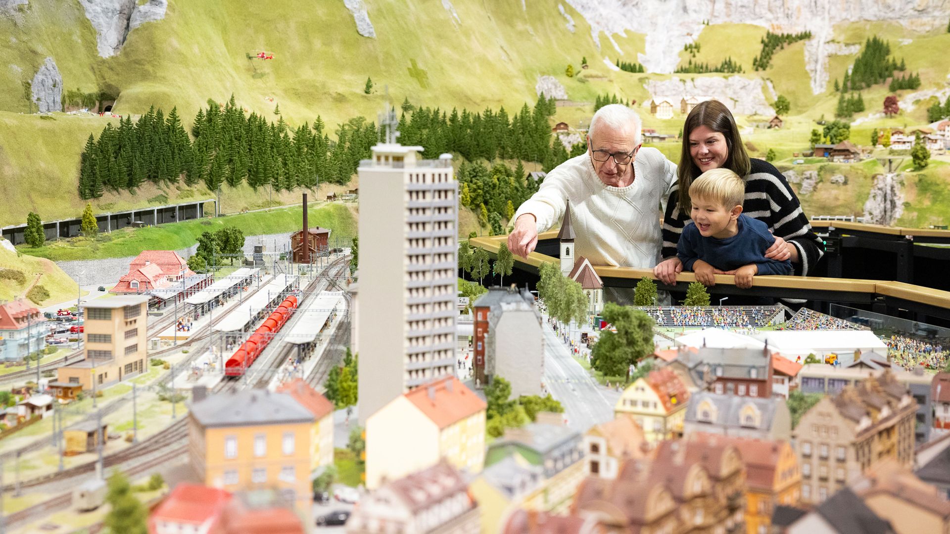 Plusieurs personnes regardent un monde miniature détaillé avec des bâtiments, des trains et des montagnes en arrière-plan