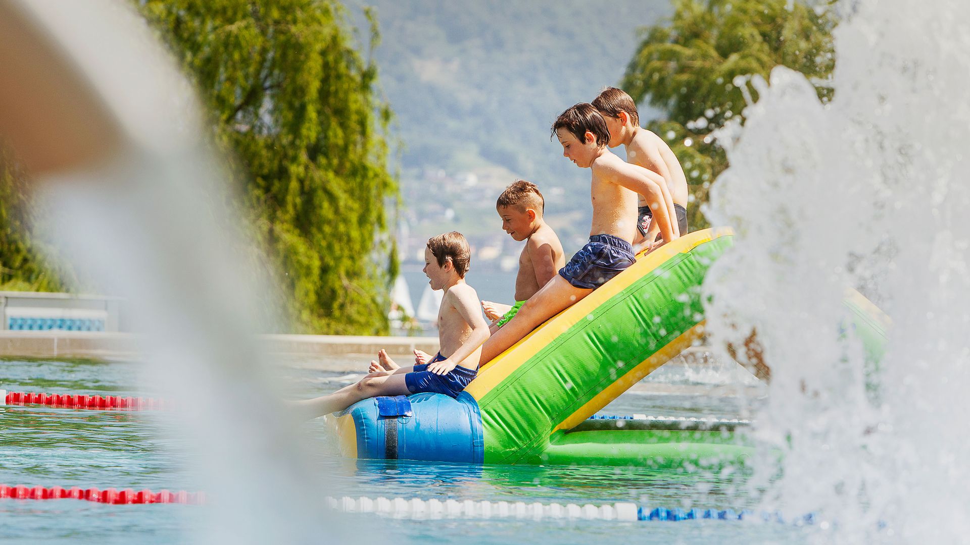 Quattro bambini seduti su uno scivolo gonfiabile nella piscina del Lido Locarno, con alberi e montagne sullo sfondo.