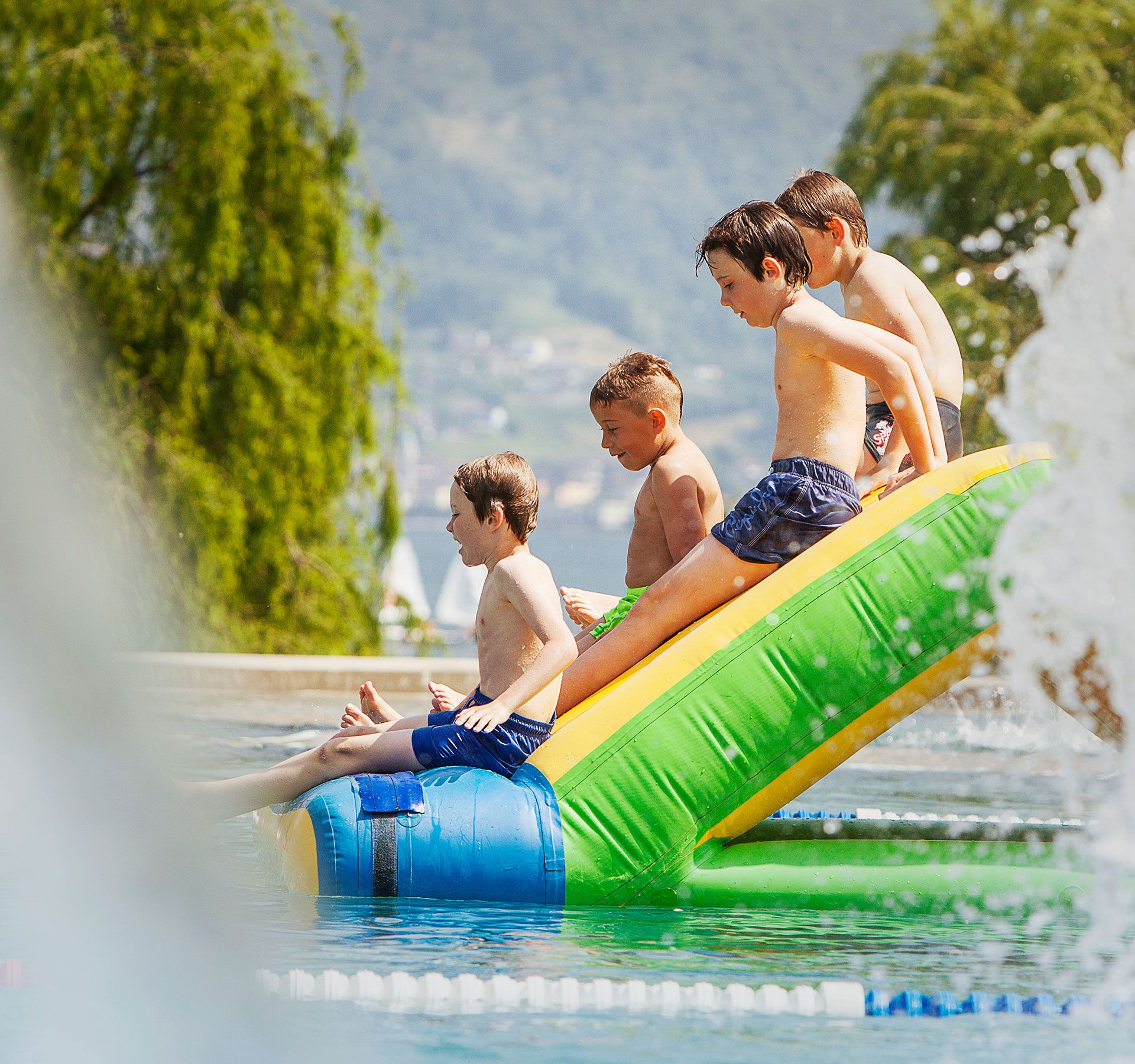 Quattro bambini seduti su uno scivolo gonfiabile nella piscina del Lido Locarno, con alberi e montagne sullo sfondo.