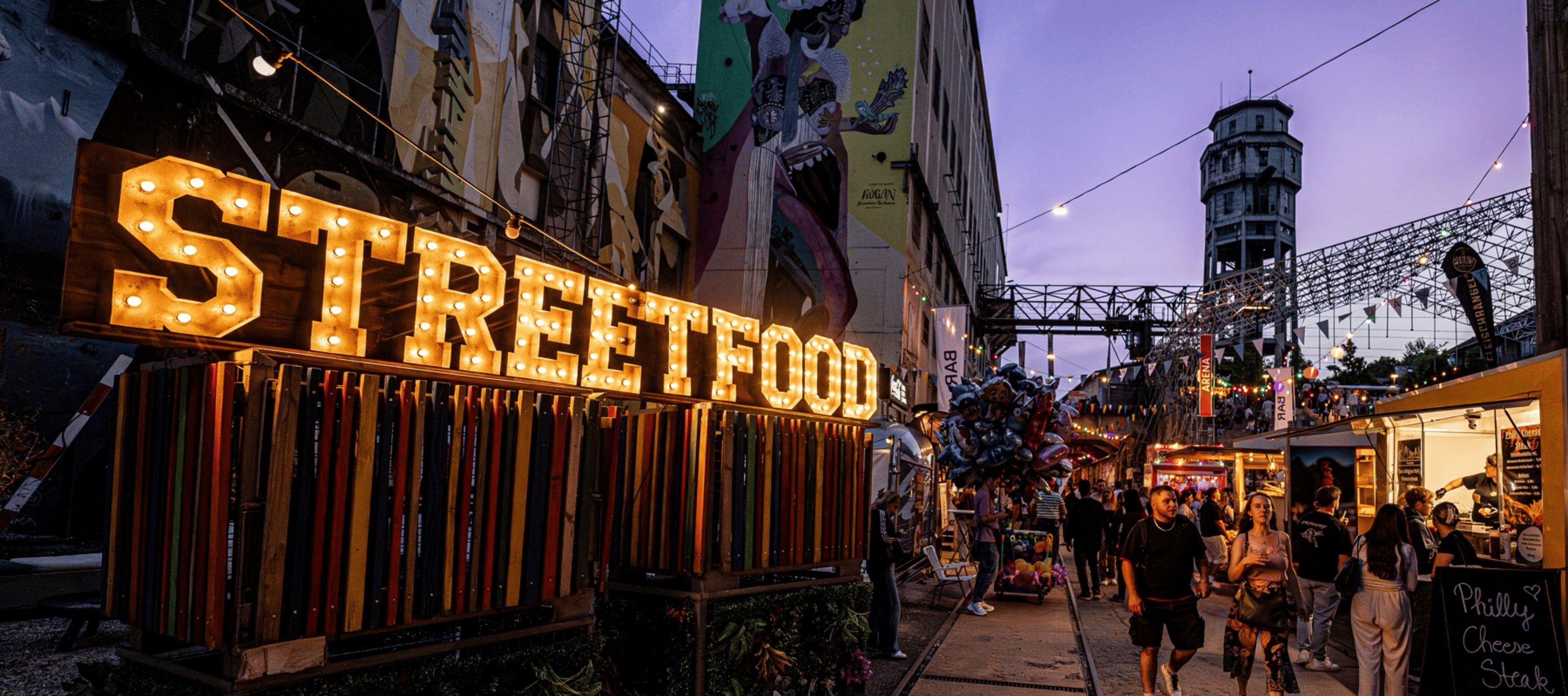Besucher schlendern über ein Streetfood-Festival mit bunten Foodtrucks, Lichterketten und Essensständen aus aller Welt.