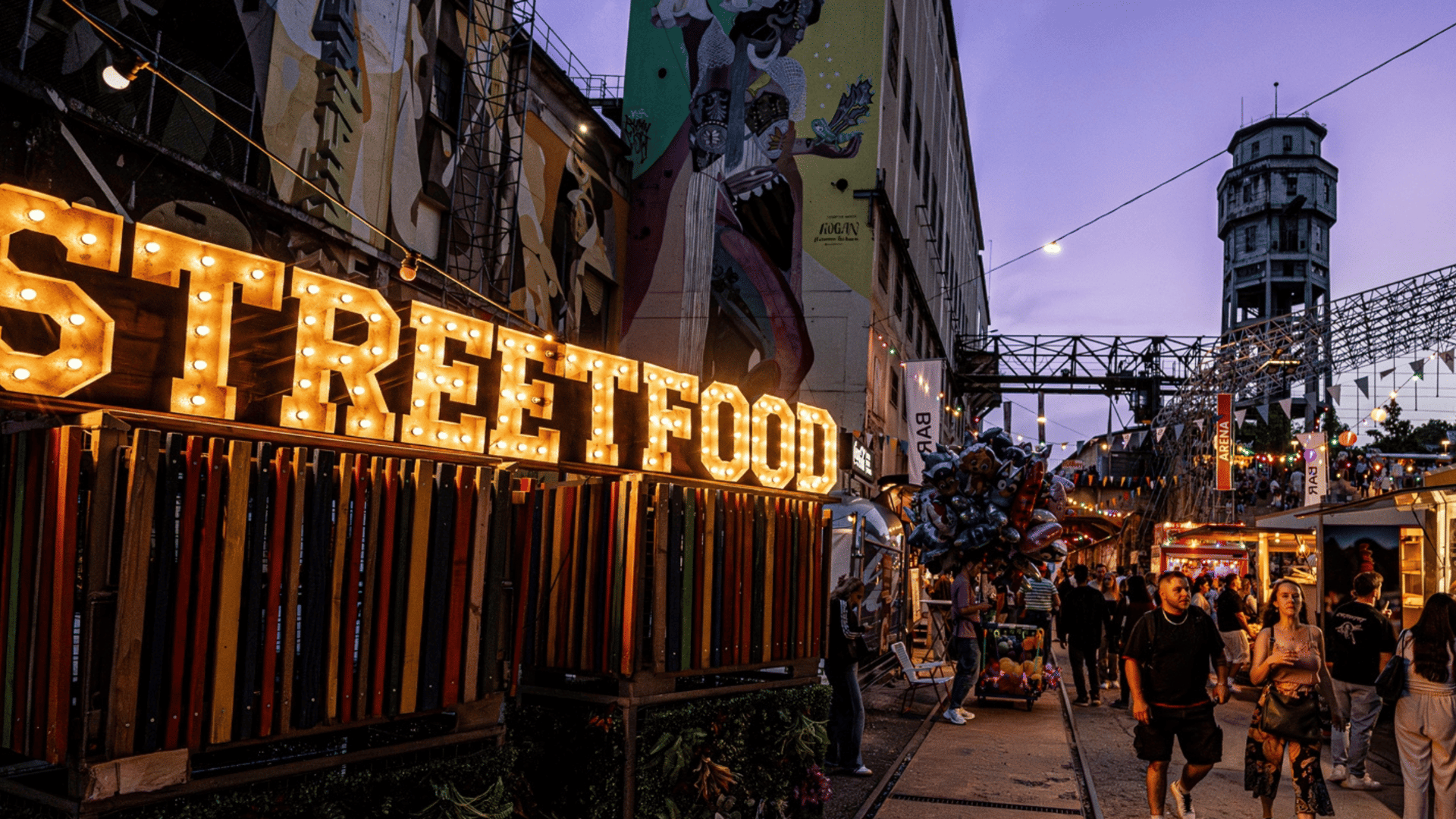 Besucher schlendern über ein Streetfood-Festival mit bunten Foodtrucks, Lichterketten und Essensständen aus aller Welt.