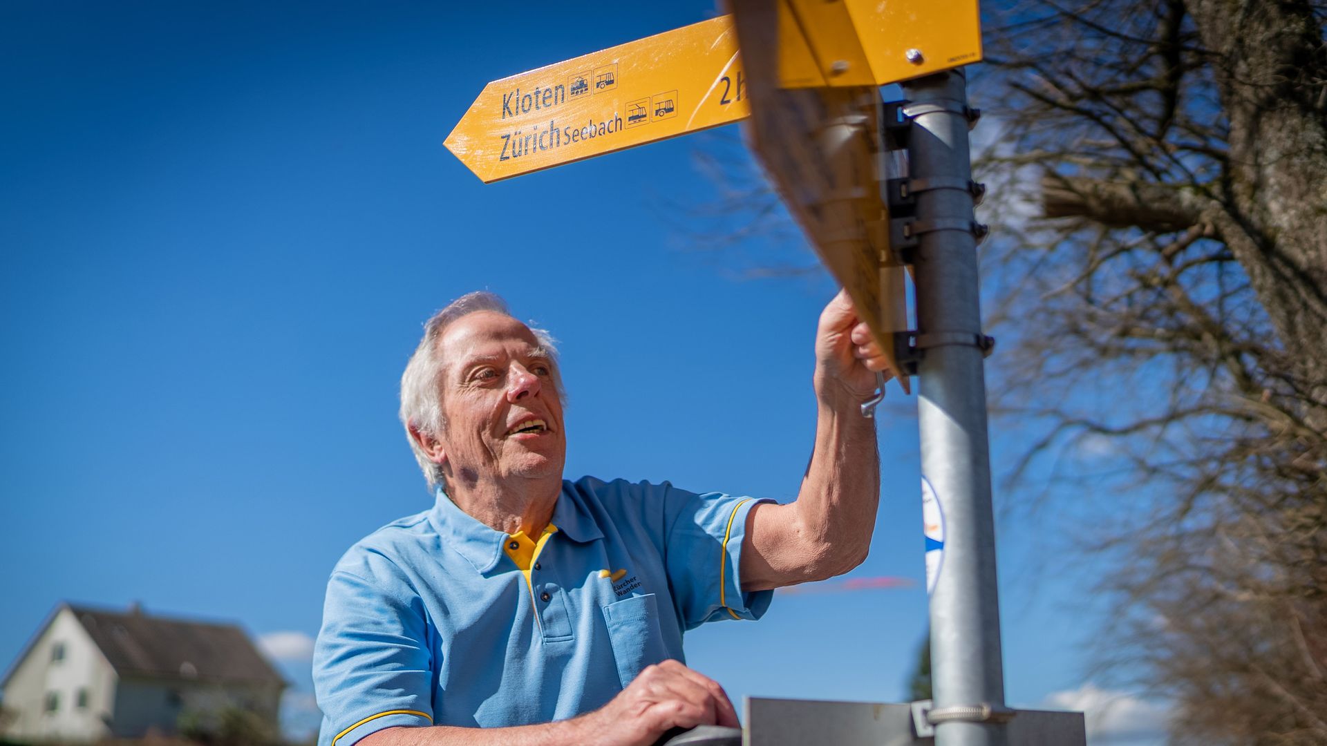 Ein Wanderer steht an einem Wegweiserschild für Wanderwege und schaut die vor sich liegende Wanderstrecke an. Das Wetter ist wunderbar sonnig, mit blauem Himmel.